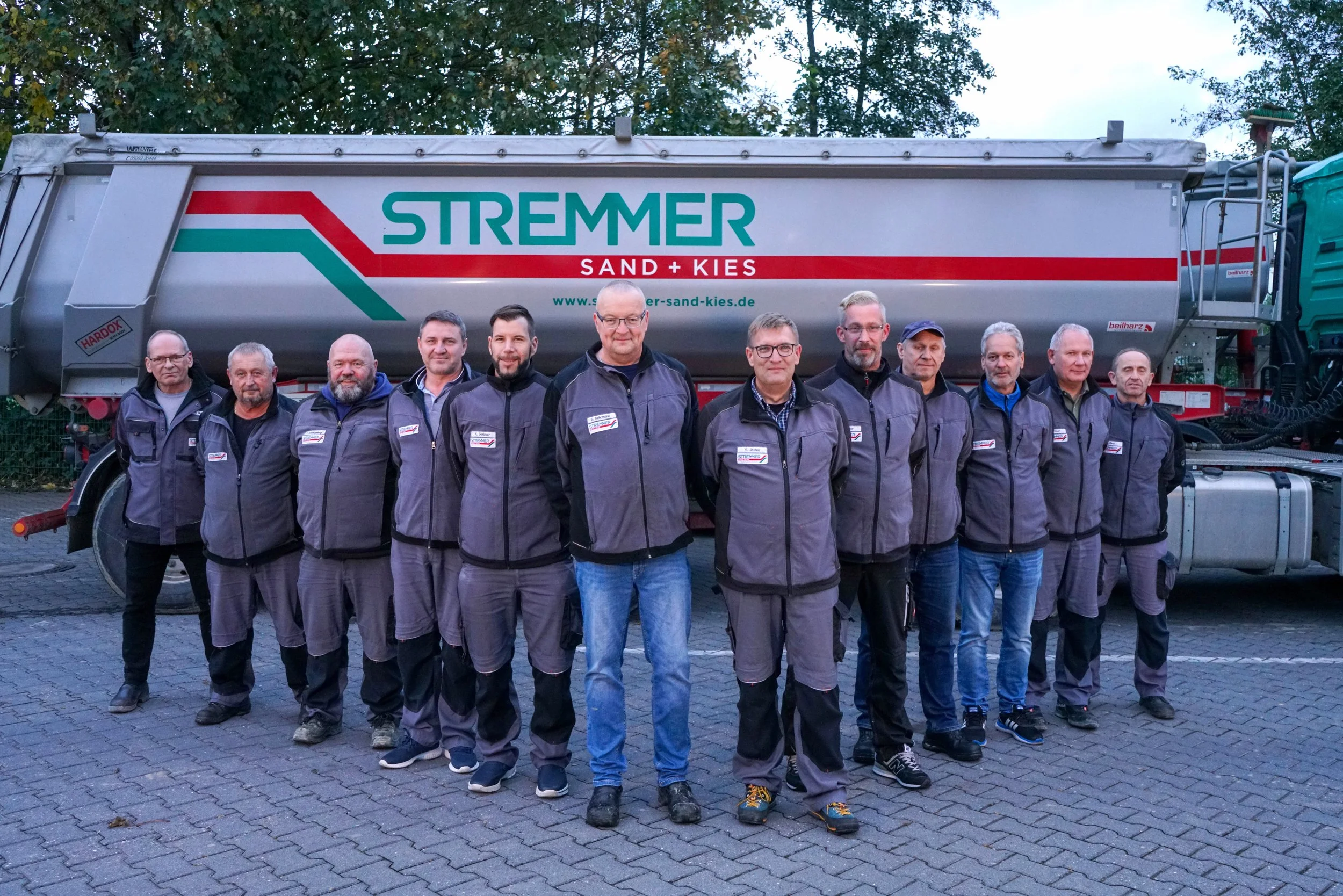 Gruppe von zwölf Männern in grauen Jacken stehen vor einem großen Silos. Das Silo trägt das Logo "STREMMER Sand + Kies" und eine Webseite. Die Männer posieren für das Foto, während im Hintergrund Bäume sichtbar sind.