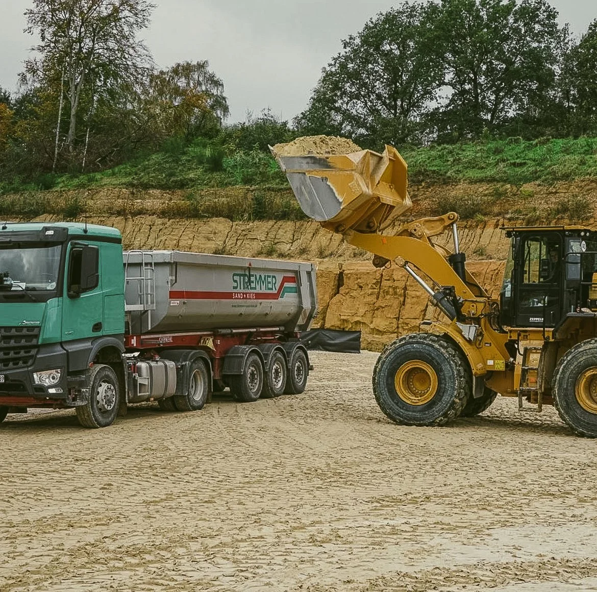 Ein Bagger hebt Sand in einen grün-blauen Lastwagen bei einer Baustelle mit Bauland und Bäumen im Hintergrund.
