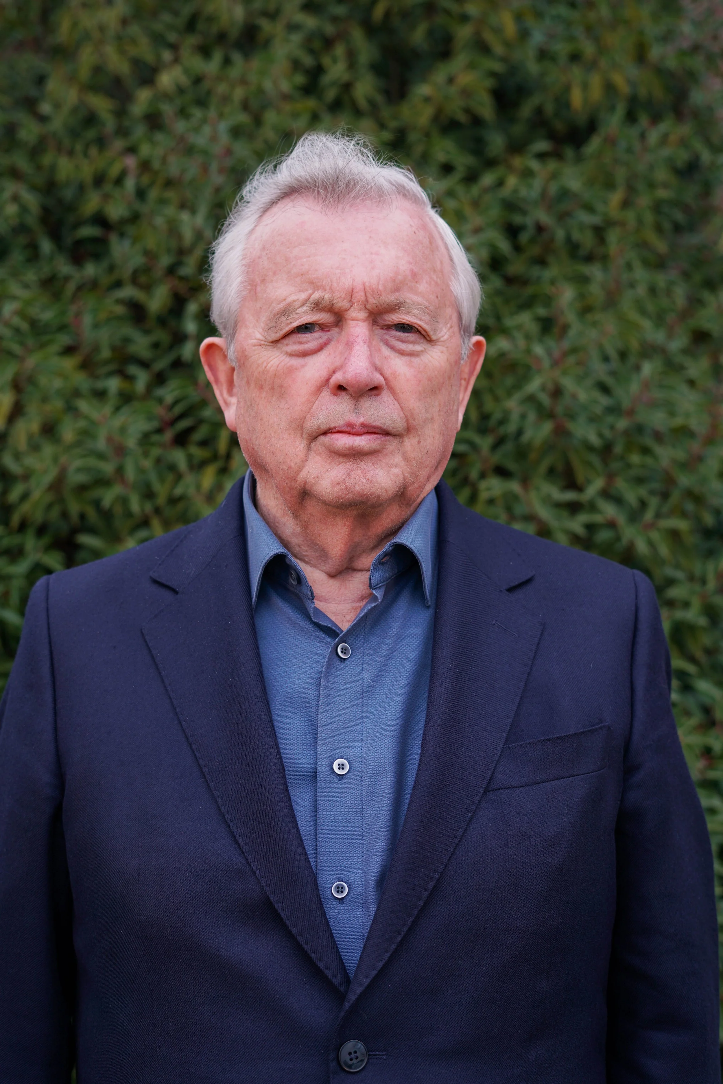 Alter Mann in Geschäftsanzug vor grünem Busch. Nahaufnahme, focus auf Gesicht und Oberkörper.