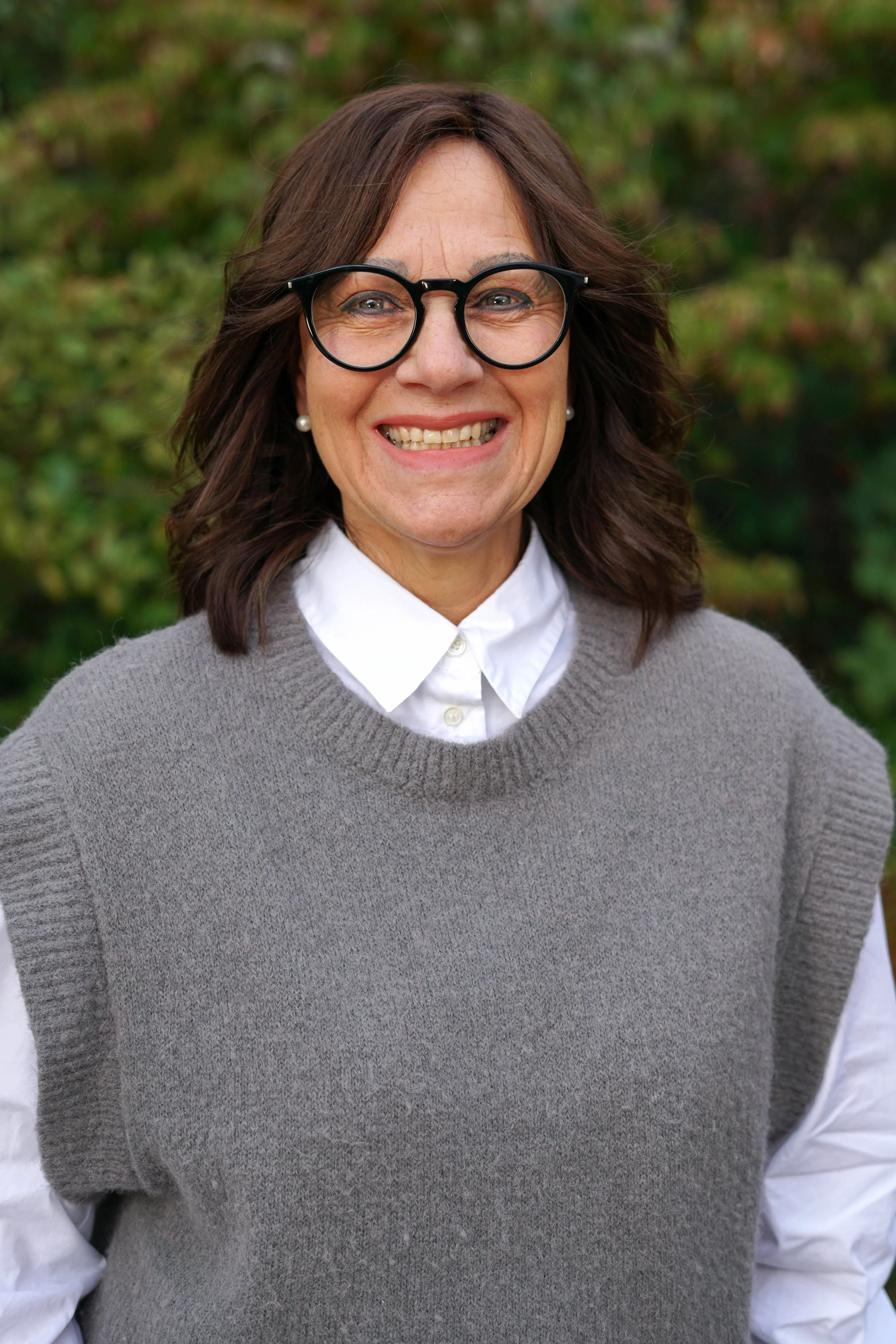 Nahaufnahme einer lächelnden Frau mit Brille, weißem Hemd und graumerwärmtem Pullover, im Freien mit grüner, unscharfer Natur im Hintergrund.