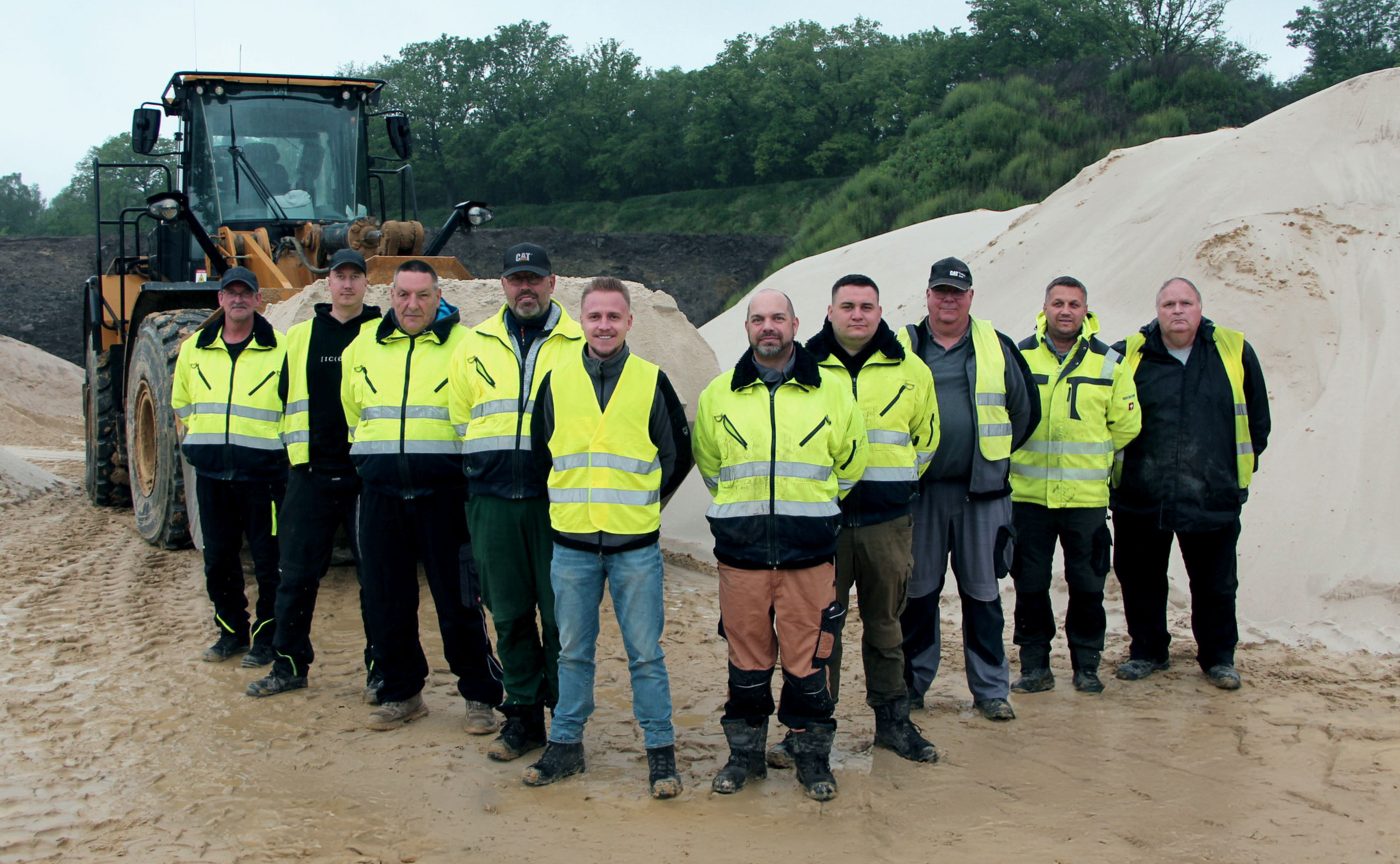 Gruppenfoto von acht Arbeitern in hoher Sichtschutzkleidung vor einem Sandberg auf einer Baustelle.