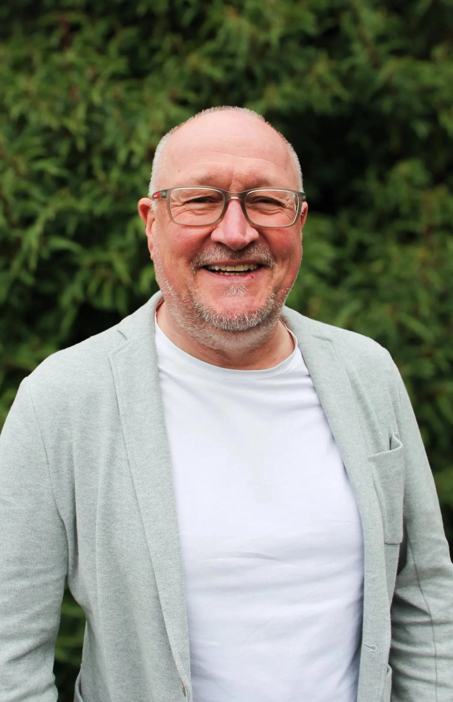 Lächelnder Mann mit Brille, grauem Blazer und weißem T-Shirt vor grüner Hecke.