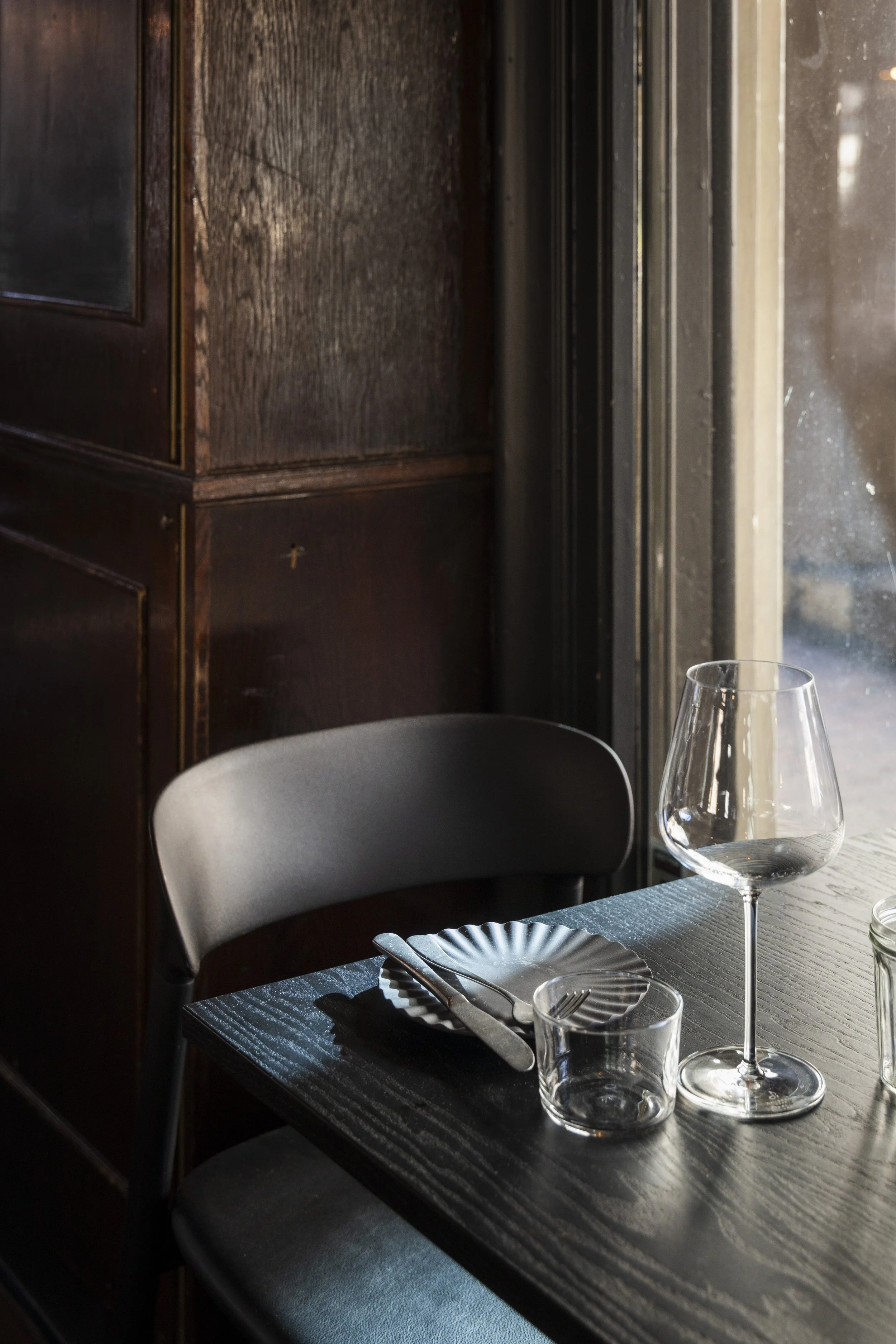 Innendørs restaurantbord med tomt vinglass, vannkjøler, kniv, gaffel, tallerken, og en grå stol. Bakgrunn med mørk trebord og vindu.