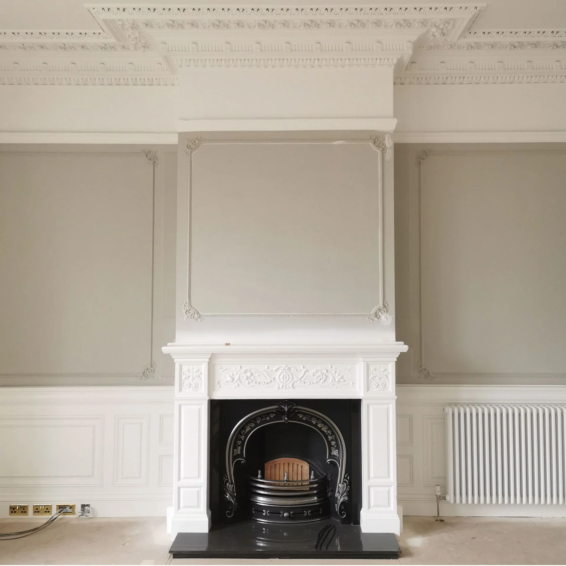 A white ornate fireplace with black accents, decorative molding, and a wooden fire grate, set in a room with white paneled walls, decorative crown molding, and a radiator on the right.