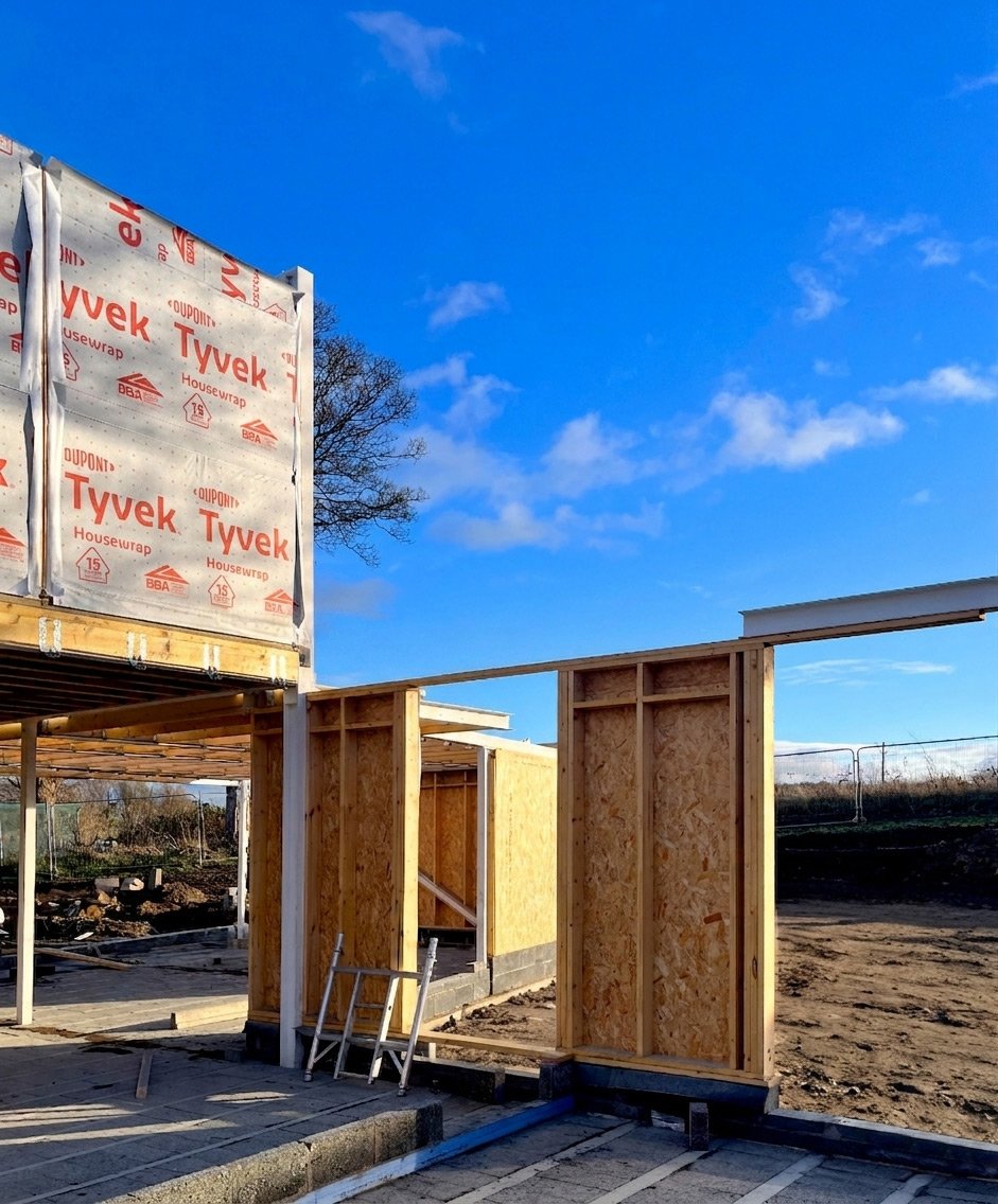 Construction site with wooden framing, a ladder, and Tyvek house wrap under a blue sky with some clouds.