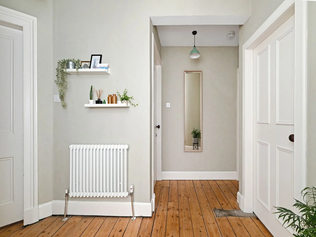 Interior view of a hallway with light-colored walls, wooden flooring, white doors, wall-mounted shelves with decorative items, a radiator, a full-length mirror, and a potted plant.