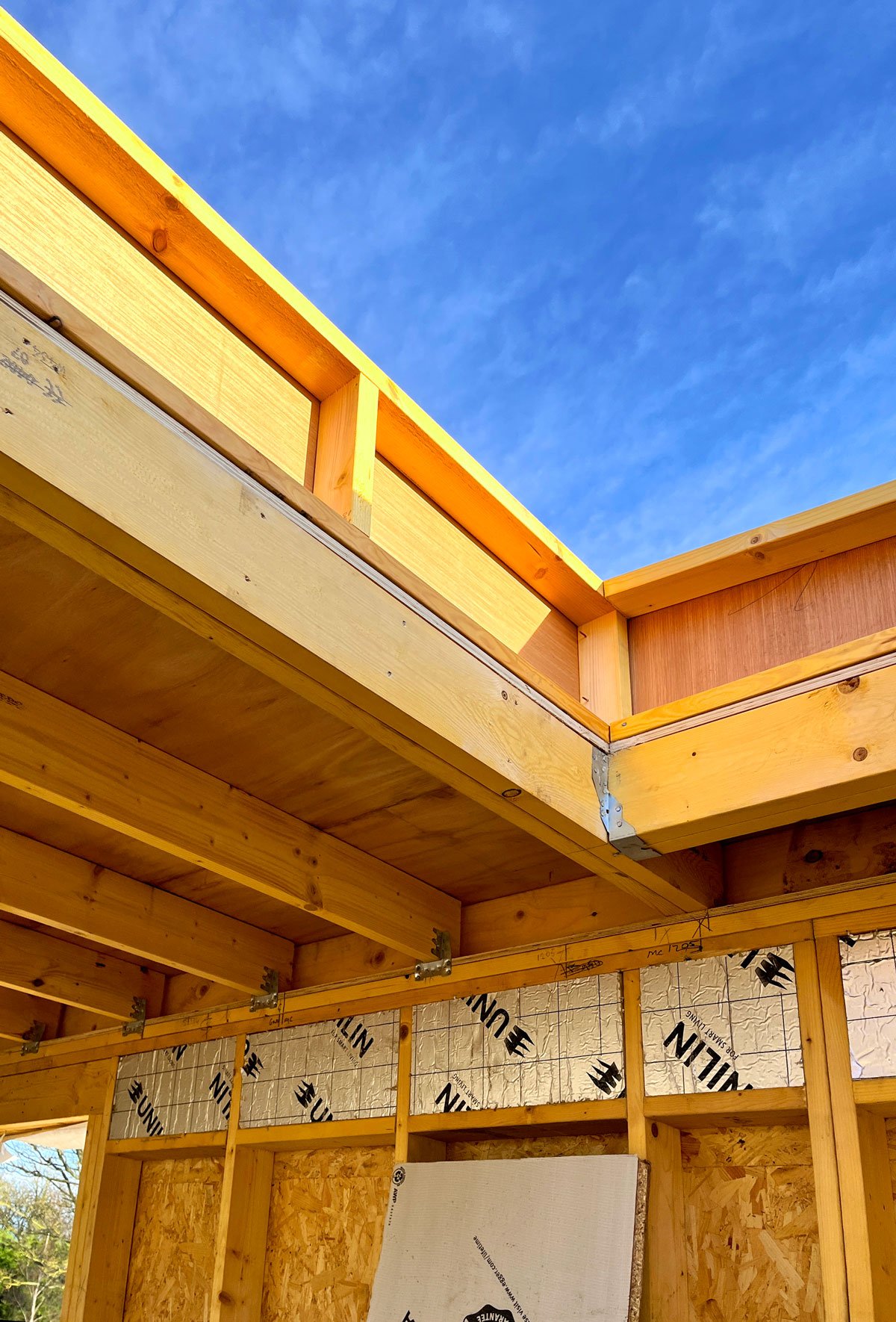 Construction site with wooden framing and insulation, showing a clear blue sky through an open roof area.