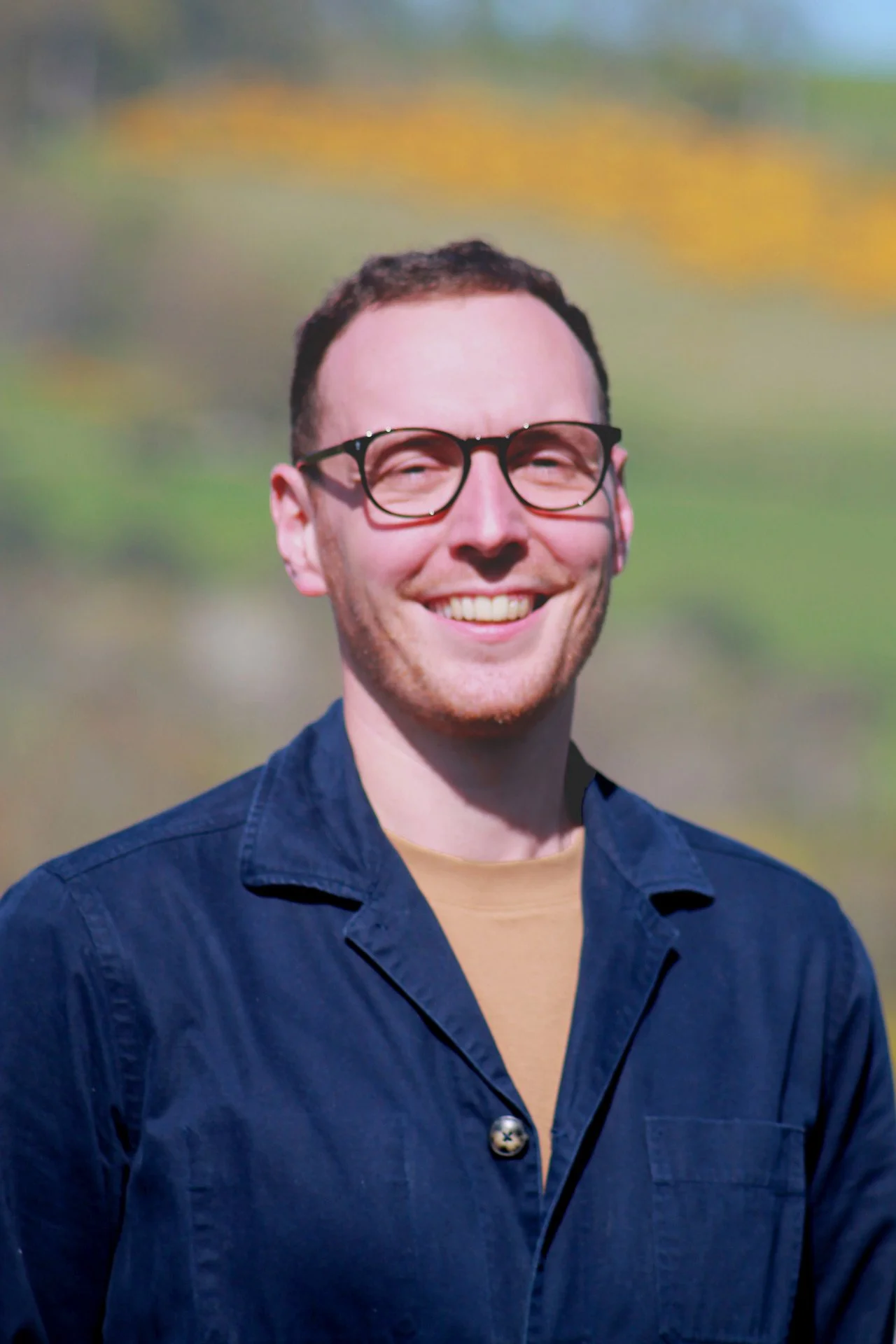 A young man with short brown hair, wearing glasses, a navy blue button-up shirt, and a beige t-shirt, smiling outdoors with a blurred natural background.
