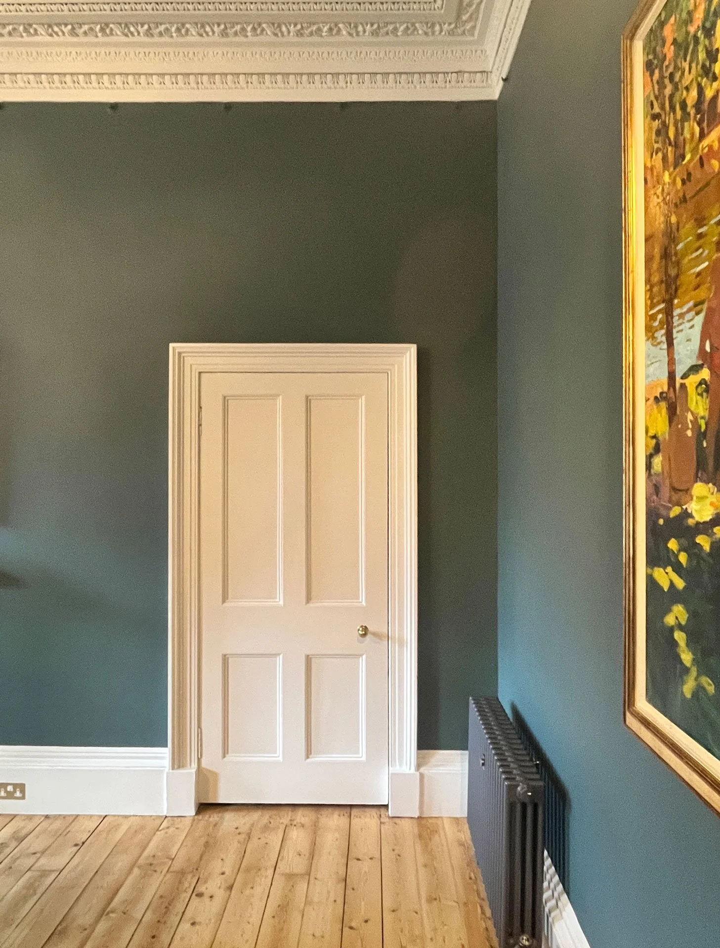 An interior room corner with a closed white door, decorated green walls, wooden flooring, a black radiator, and a large framed colorful painting on the right.