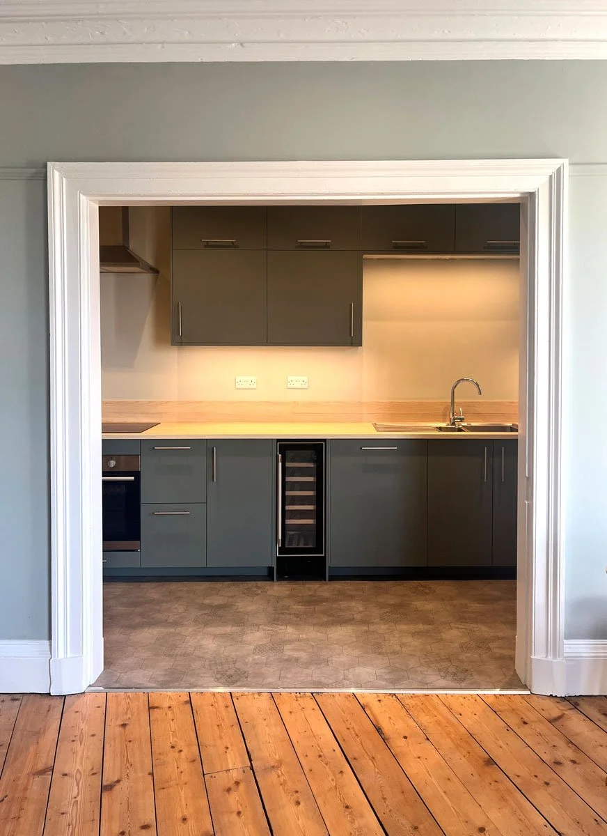 Kitchen seen through doorway with dark gray cabinets, built-in oven, wine cooler, and sink, with a light wood countertop and gray flooring.