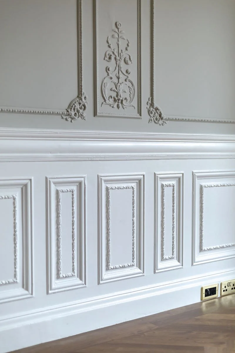 Close-up of an ornate cream-colored wall with decorative molding and paneling, with a wooden floor and electrical outlets at the bottom.