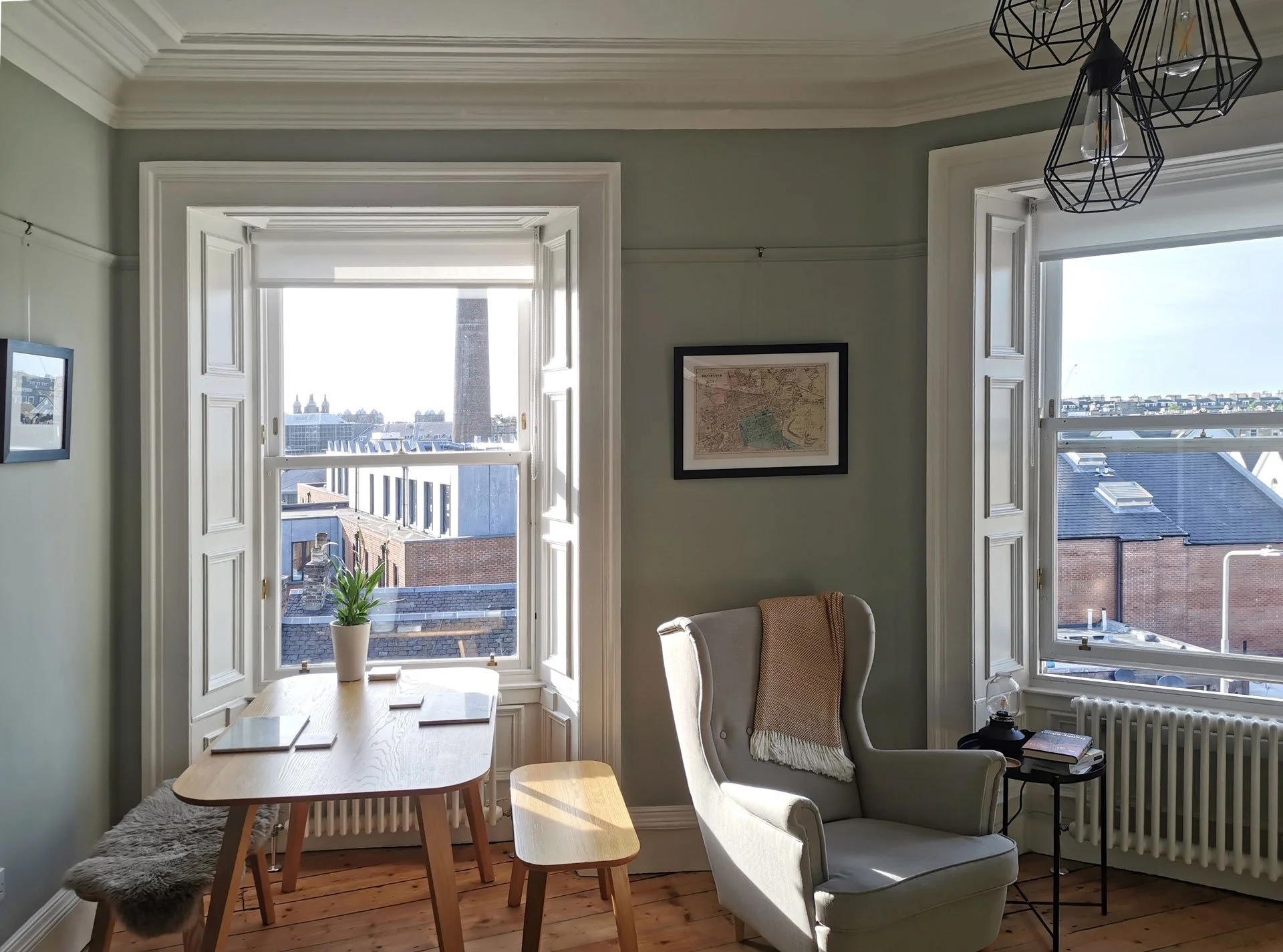 Bright room with large windows, a dining table with three chairs, a potted plant, a framed map on the wall, and a cozy armchair near a small side table holding books and a lamp, overlooking a cityscape.