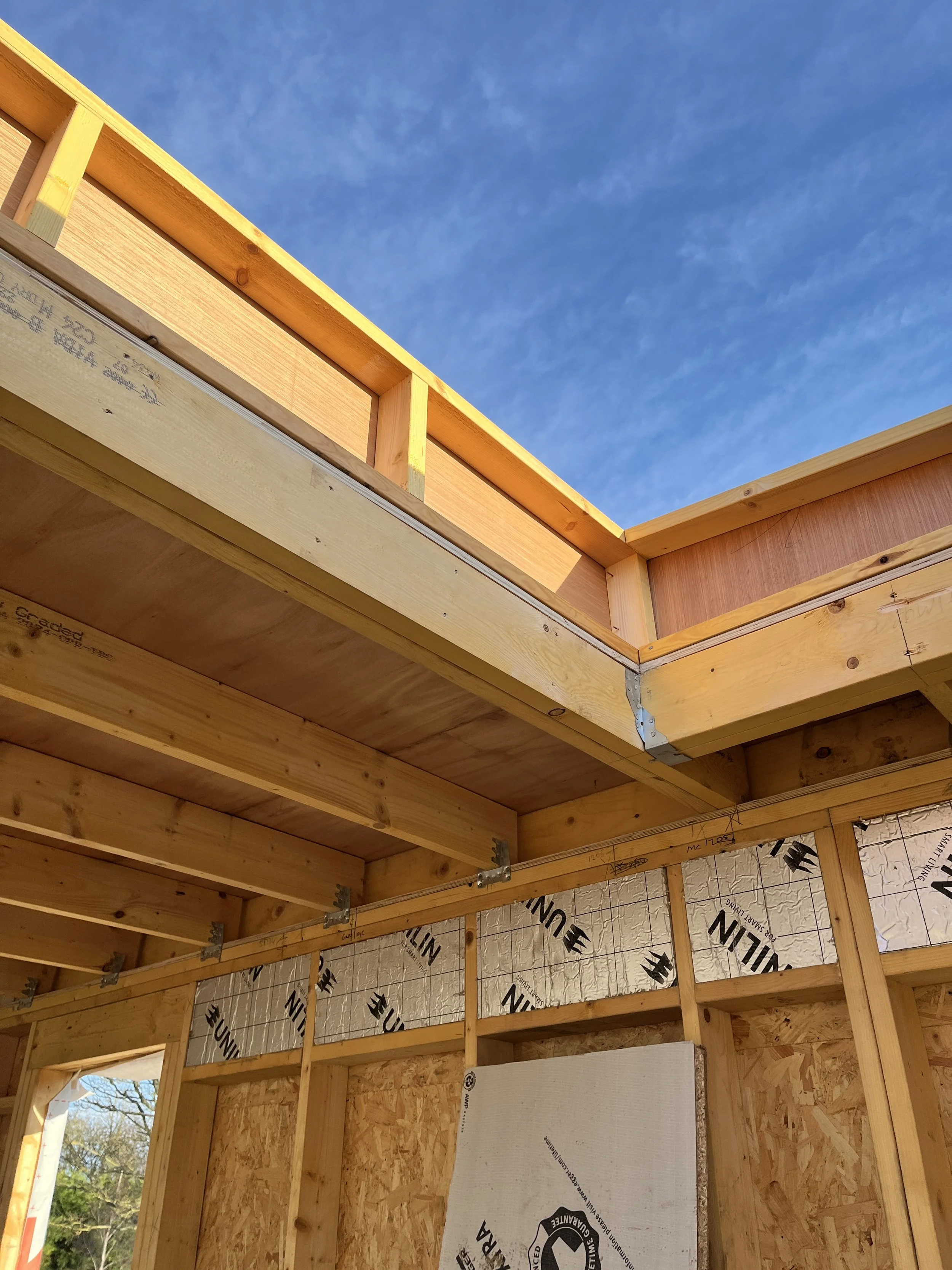 Interior view of a wooden house under construction, showing the framing and insulation, with the sky visible through an opening at the top.