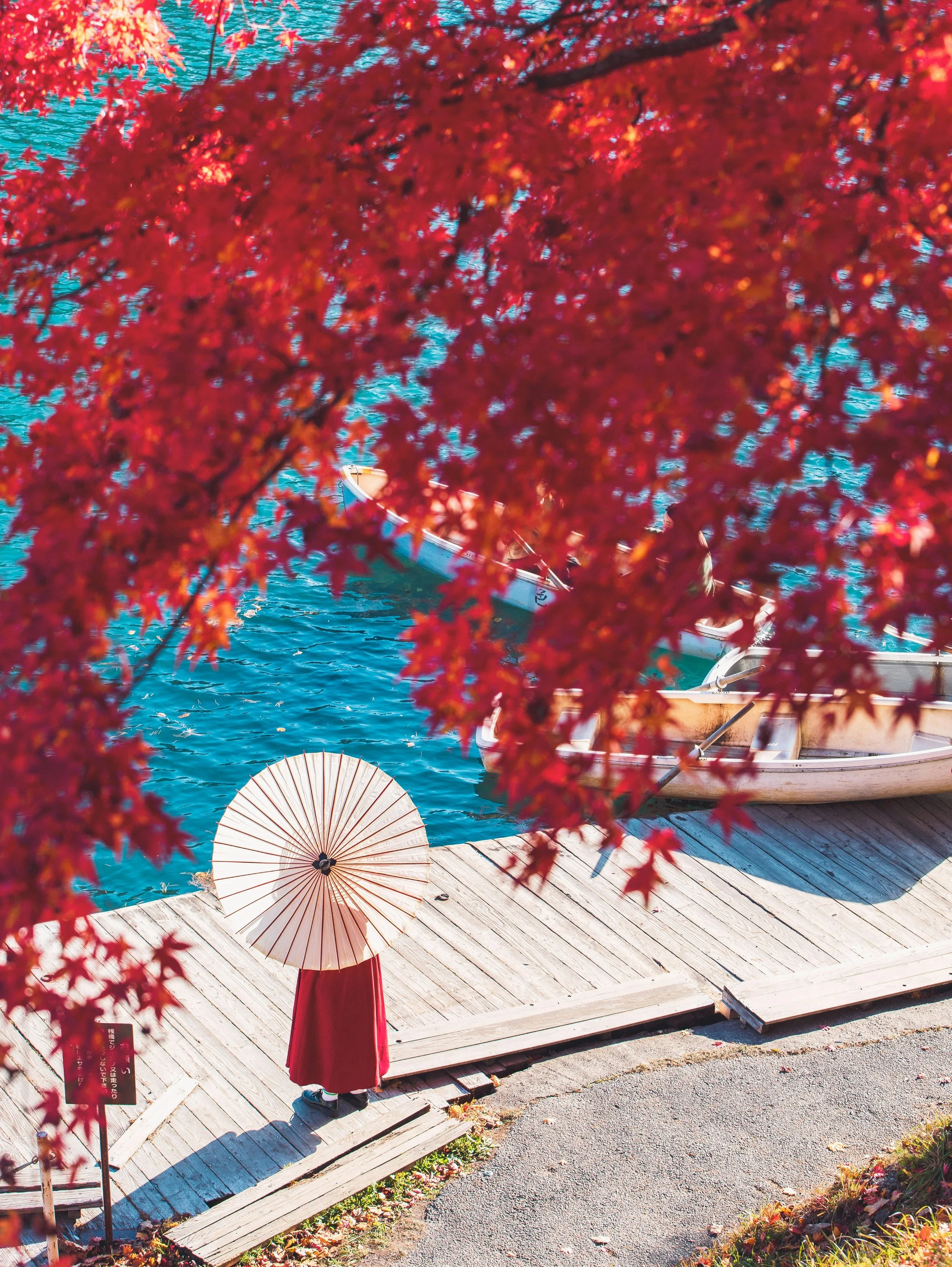 Vibrant red maple leaves in autumn framing a view of a blue lake with a red parasol and wooden deck
