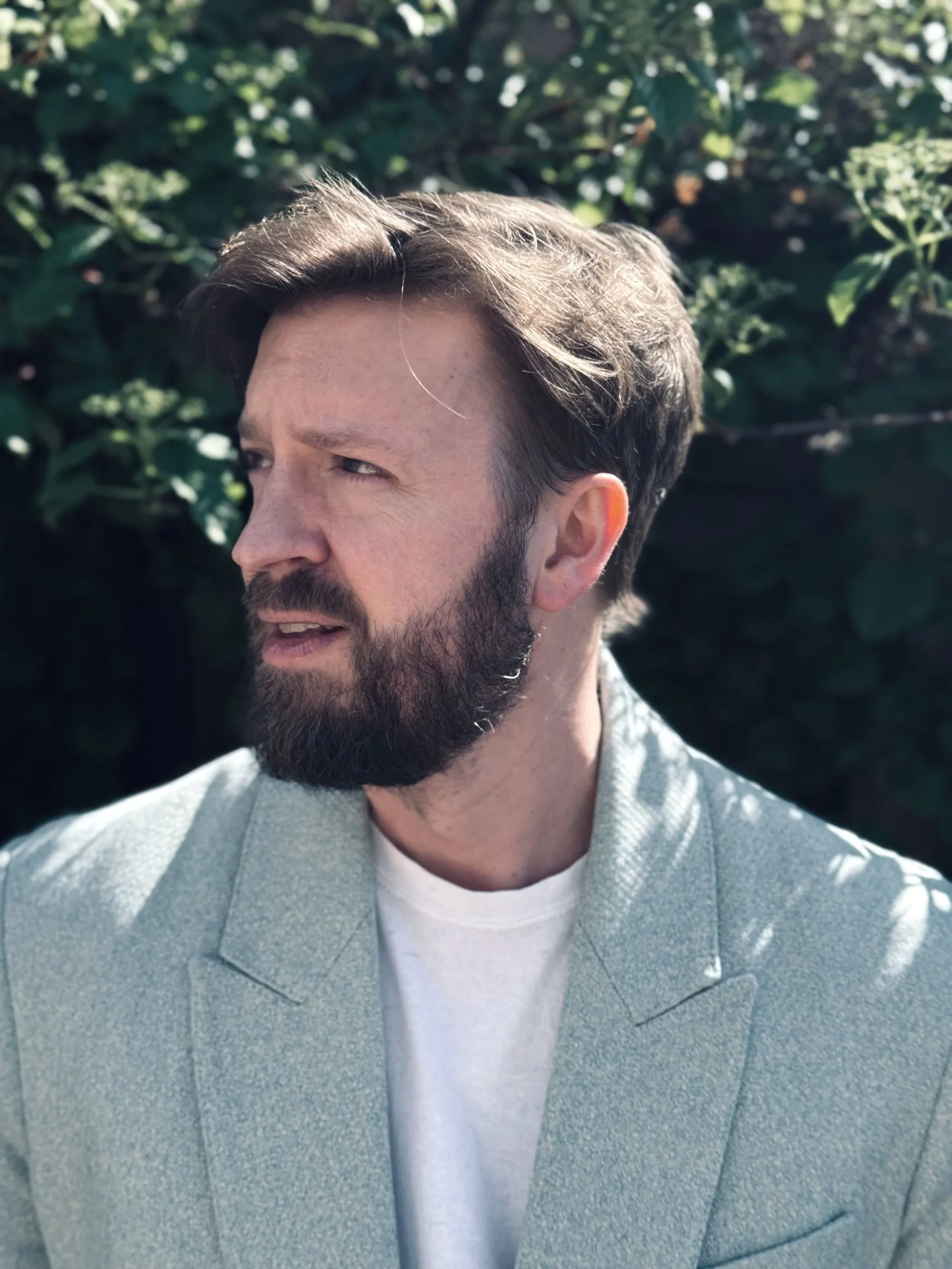 A man with a beard and mustache looking to his right, wearing a light grey blazer and a white t-shirt, outdoors with green foliage in the background.