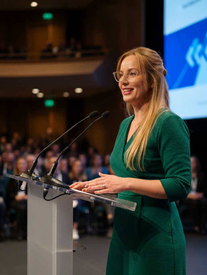 A woman with long red hair, glasses, and a green dress, speaking at a podium with microphones during a conference.