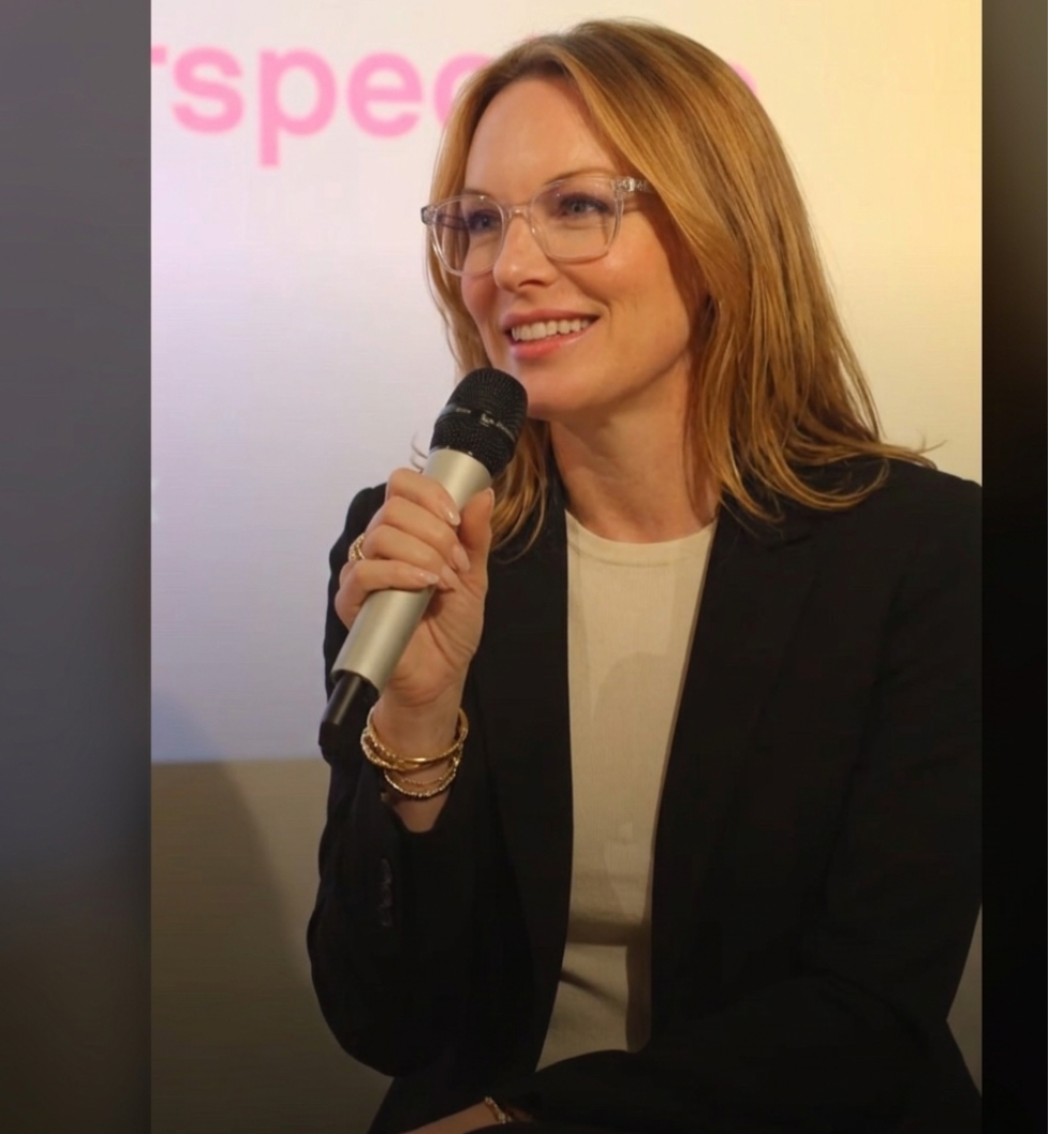 A woman with red hair and glasses holding a microphone, smiling during a presentation or speech.