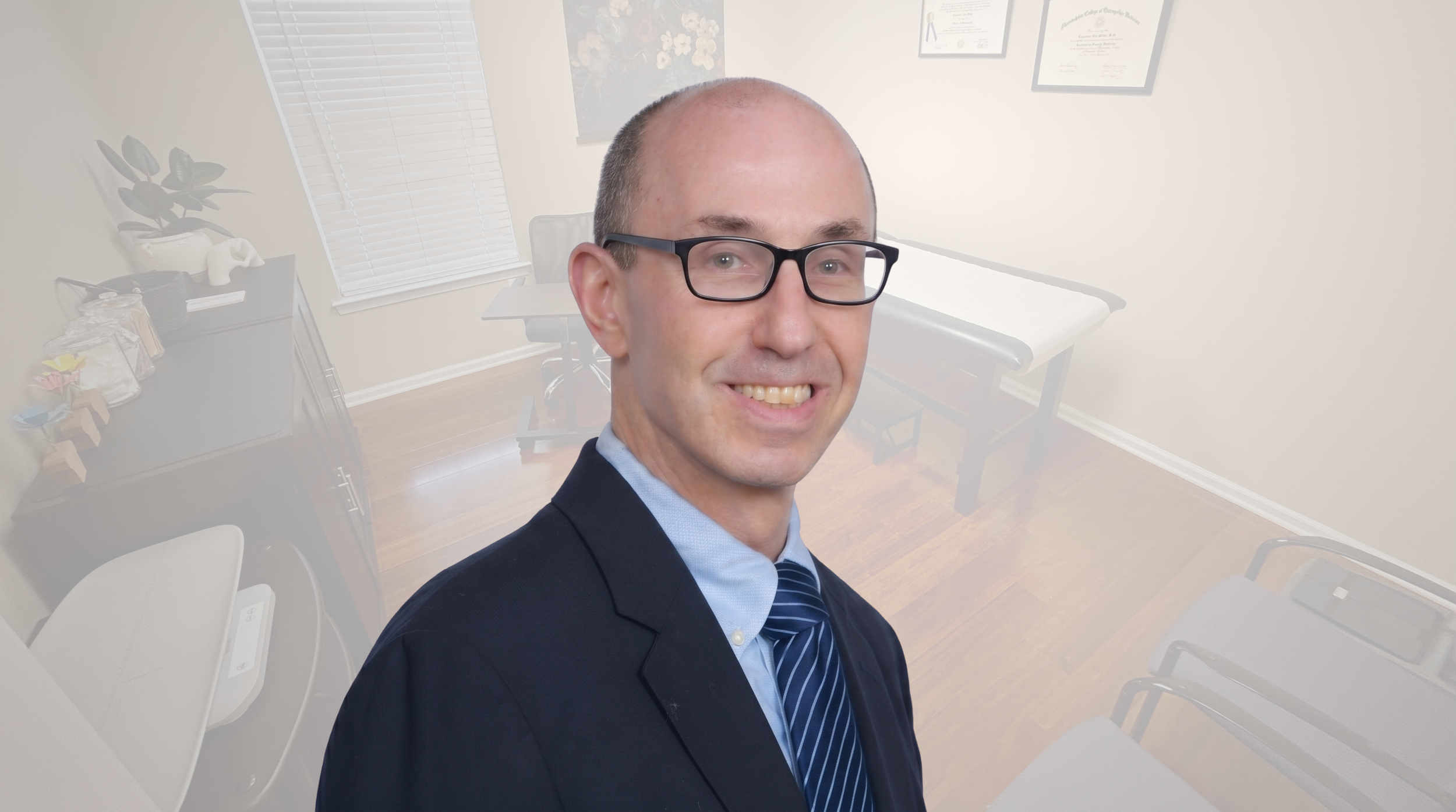 A man in business attire smiling in an office with a table, framed diplomas on the wall, and a potted plant.