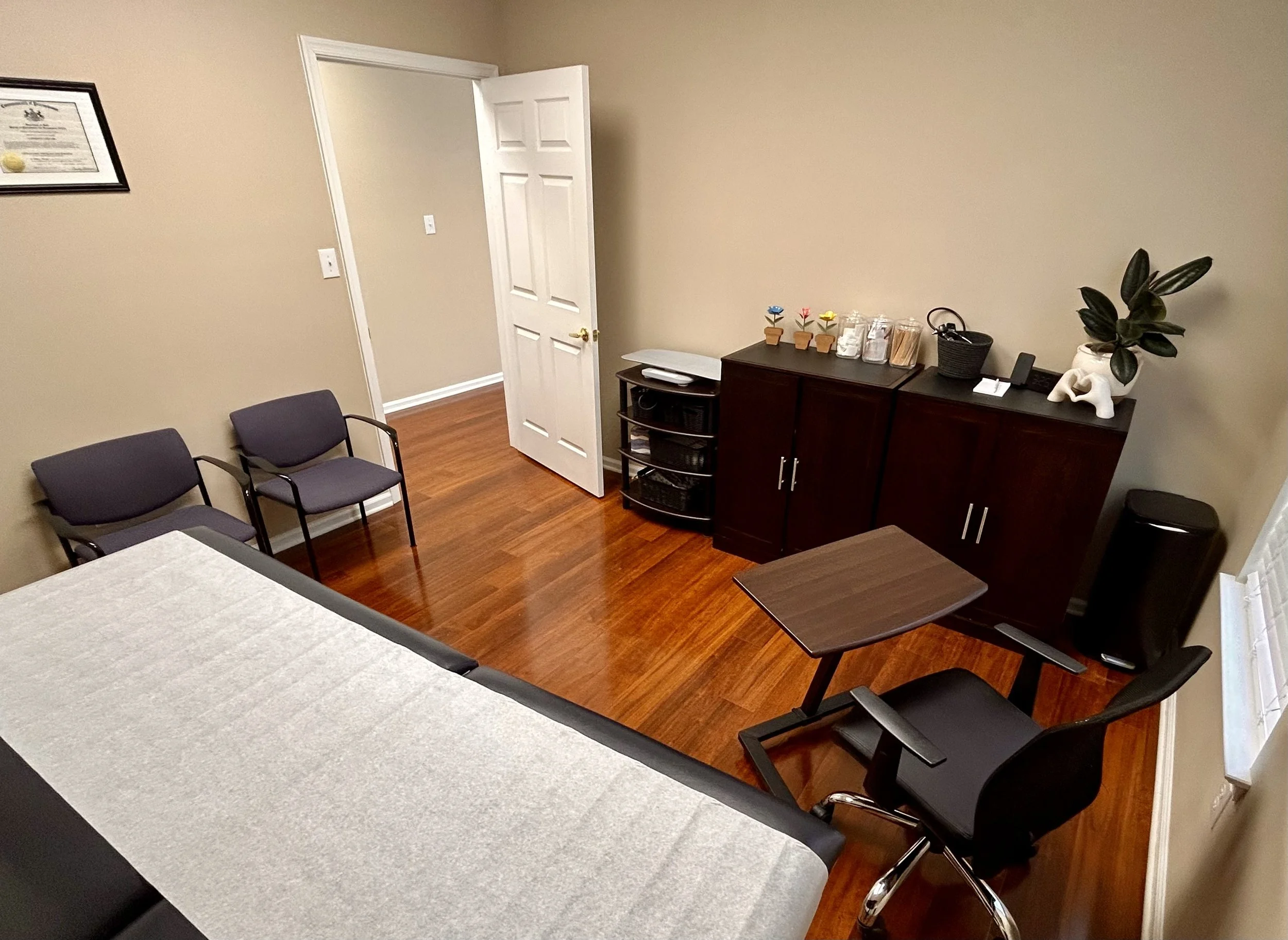 Office room with beige walls, dark brown furniture, two chairs, a small shelving unit, a plant on a cabinet, and a framed certificate on the wall.