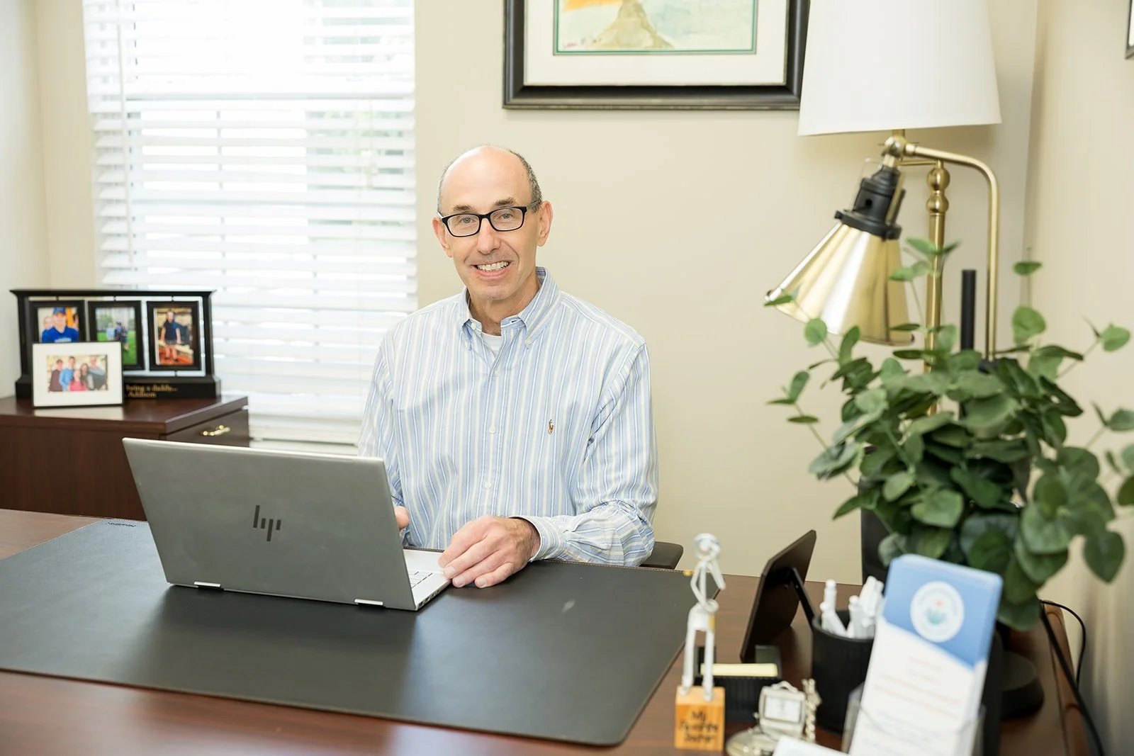 Dr. Lawrence Miller (Larry) sitting at a desk with a laptop, smile, in an office with framed photos, a plant, and a lamp.