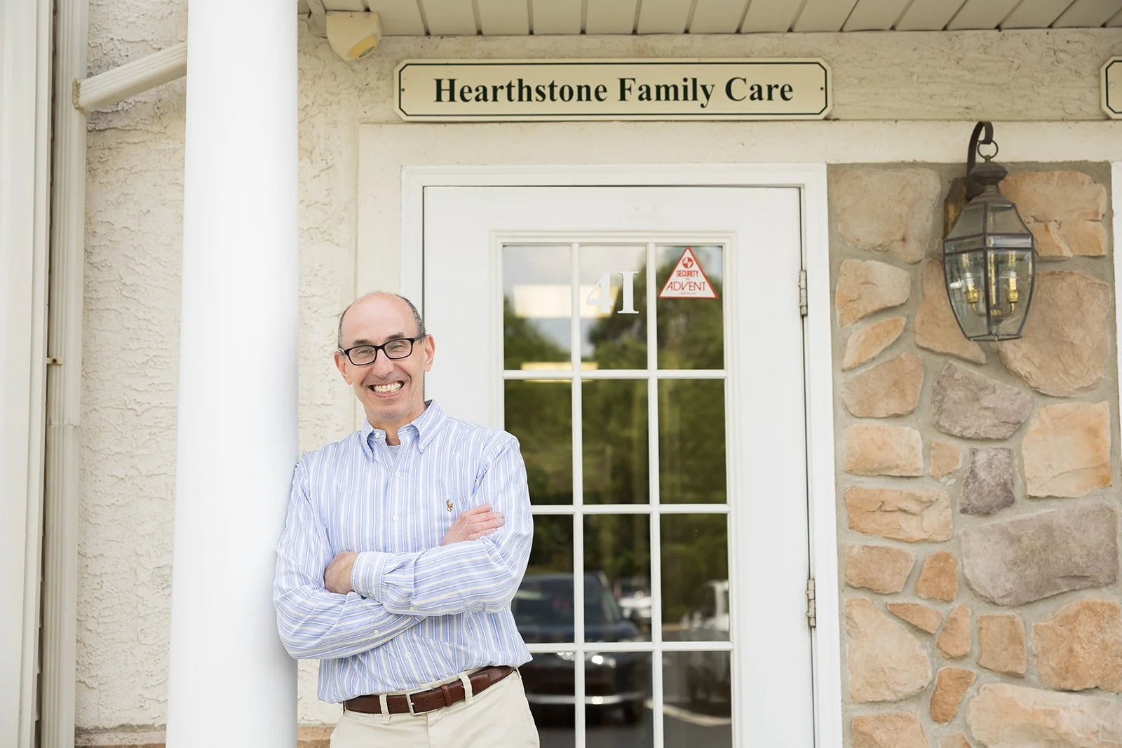 Dr. Lawrence Miller (Larry) standing outside of a building with a sign that reads "Hearthstone Family Care." He is wearing glasses, a light blue and white striped shirt, and beige pants, standing with his arms crossed.