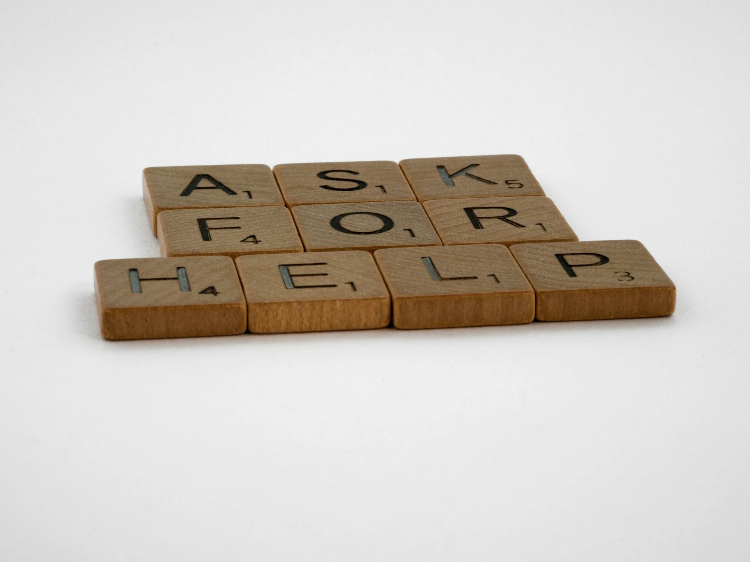 A close-up of wooden Scrabble tiles spelling out "ASK FOR HELP" on a white background.