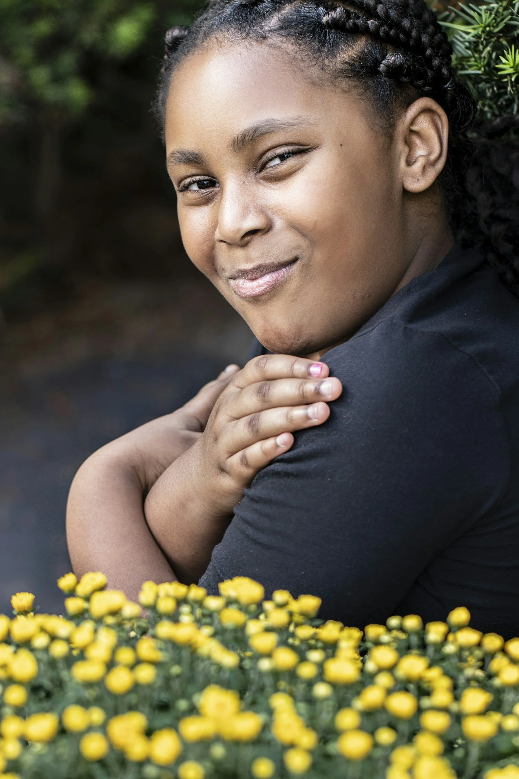 A young woman with braided hair smiling and looking at the camera, with yellow flowers in the foreground and greenery in the background.