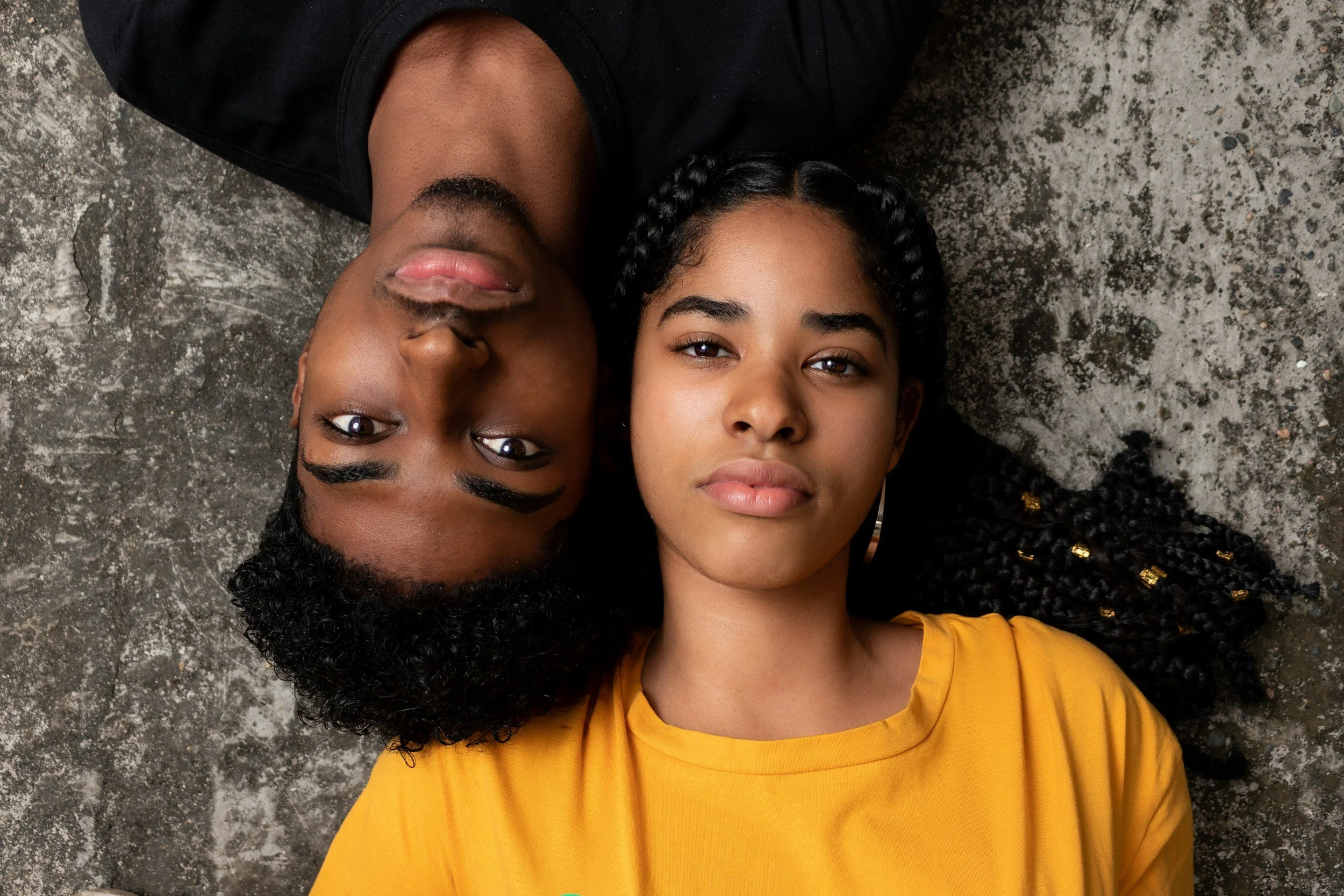 Two young women lying on a concrete floor, one with curly hair and dark skin, wearing a black top, and the other with braided hair and medium skin tone, wearing a yellow shirt, looking directly at the camera.