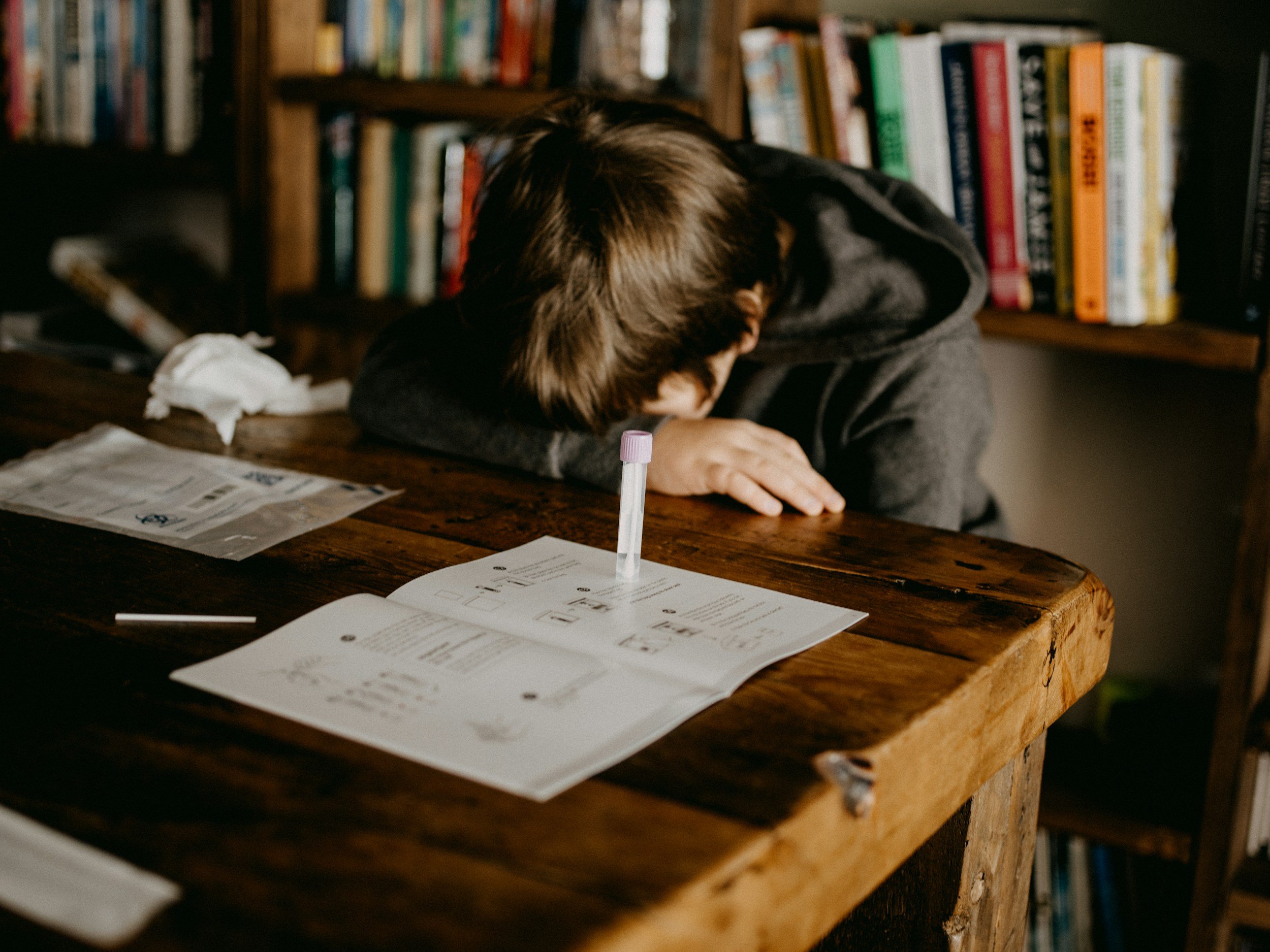 A person with brown hair and a dark jacket is resting their head on their arms on a wooden table, with a library or bookshelf in the background. There are papers and a marked test tube on the table.