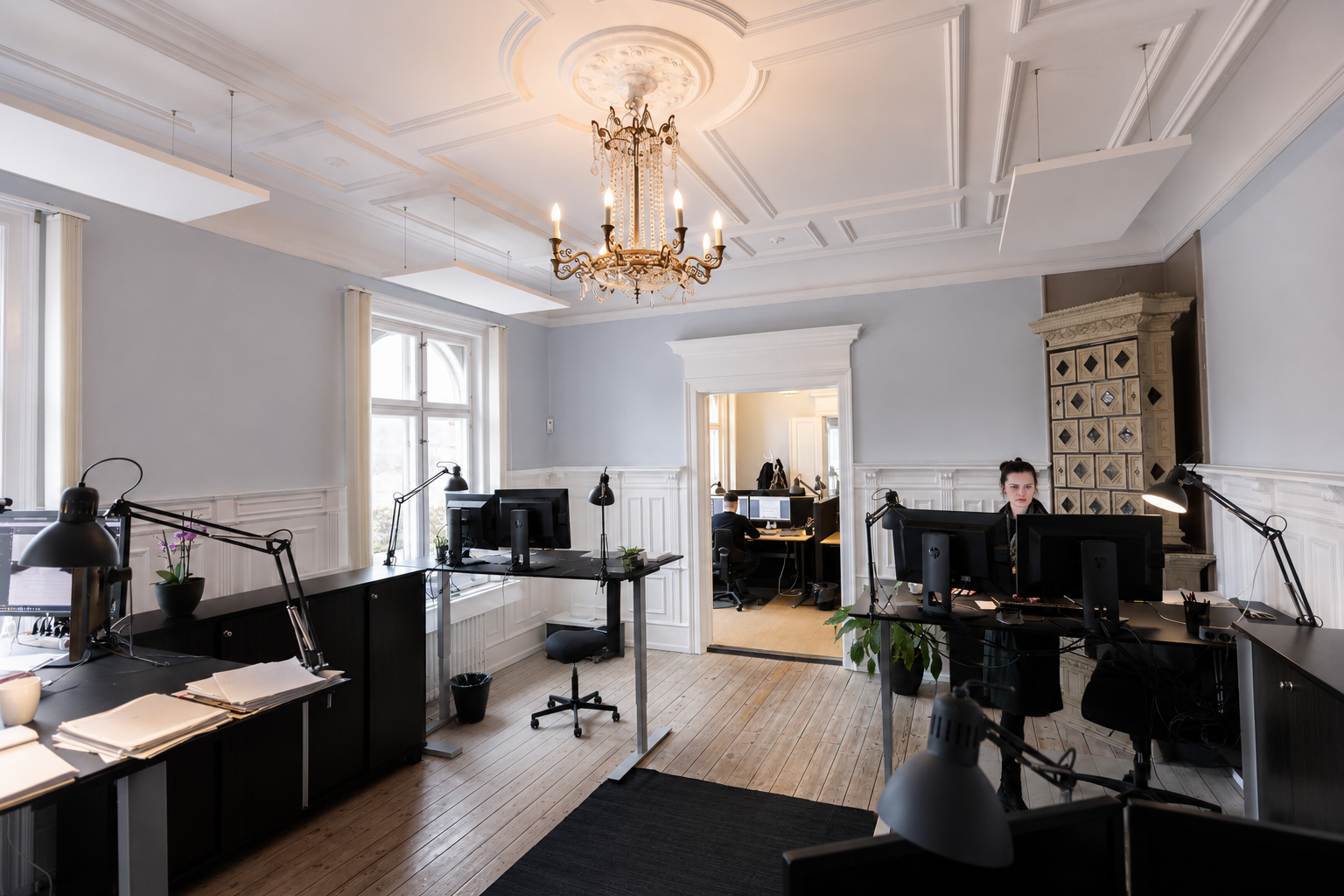 Kontor med flere arbejdsstationer, computere, skrivebordlamper og en kvinde, der sidder foran en computer. Der er store vinduer, en gammel pejs og en stor lysekrone i loftet.