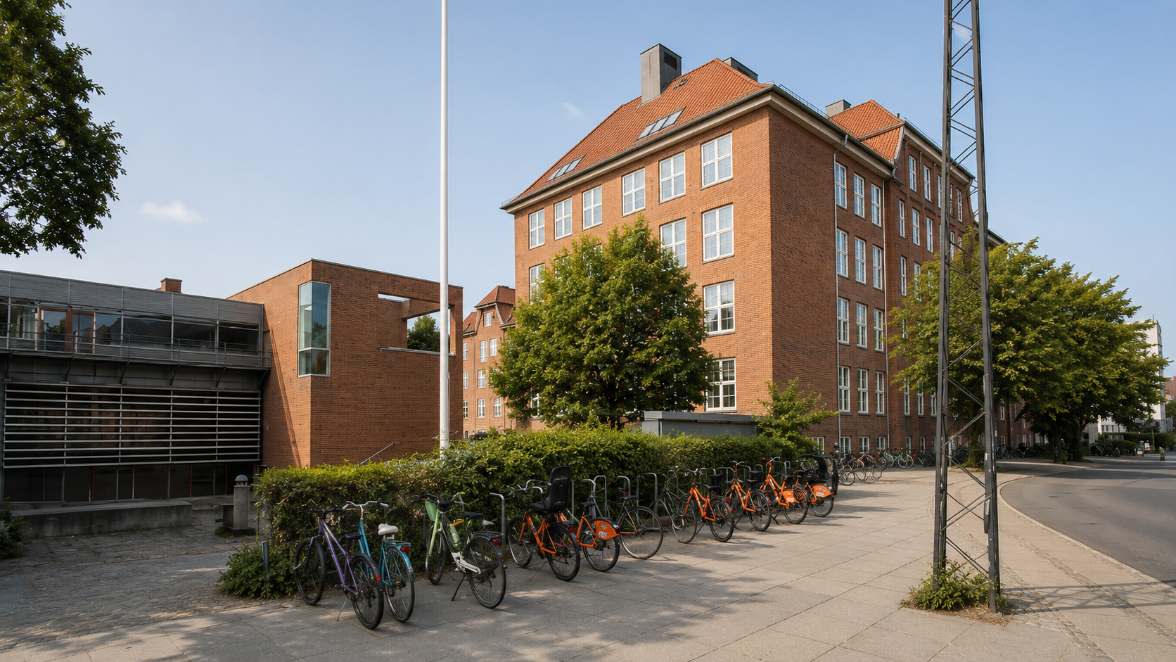 Billede af en gade med cykler parkeret foran en grøn busk, med en stor rød murstensbygning i baggrunden og nogle træer langs fortovet.