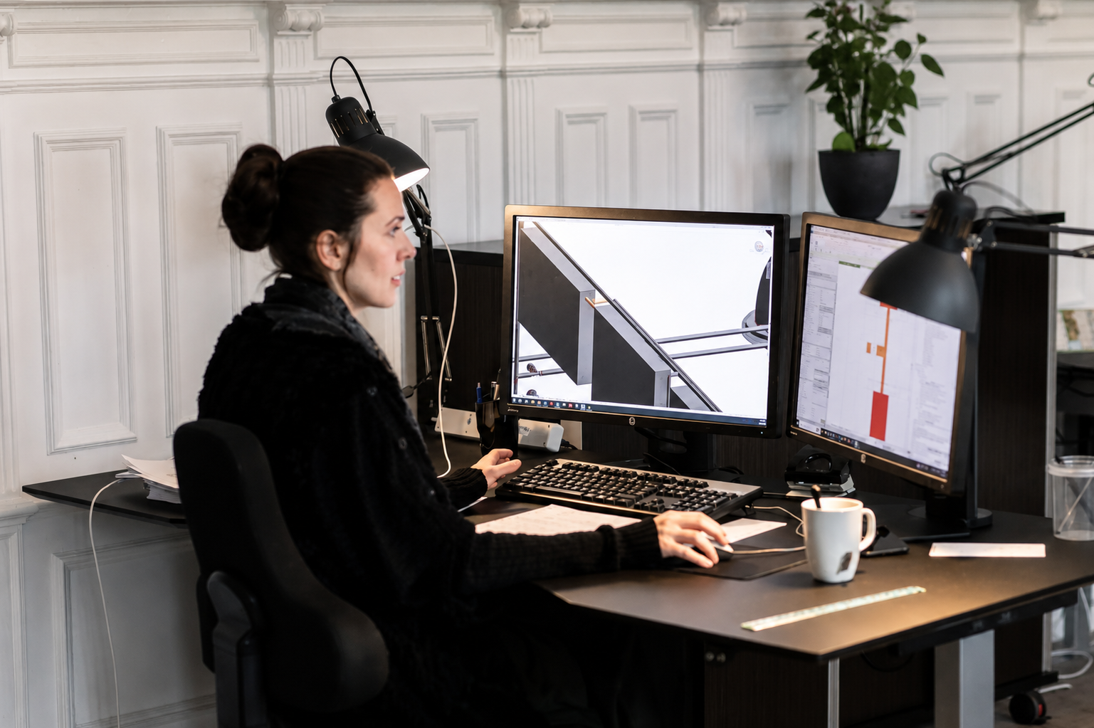Kvinde, der sidder ved en kontorbord med to computere, arbejdende på CAD- eller ingeniørsoftware, omgivet af kontorindretning, med planter og skrivebordslampe.