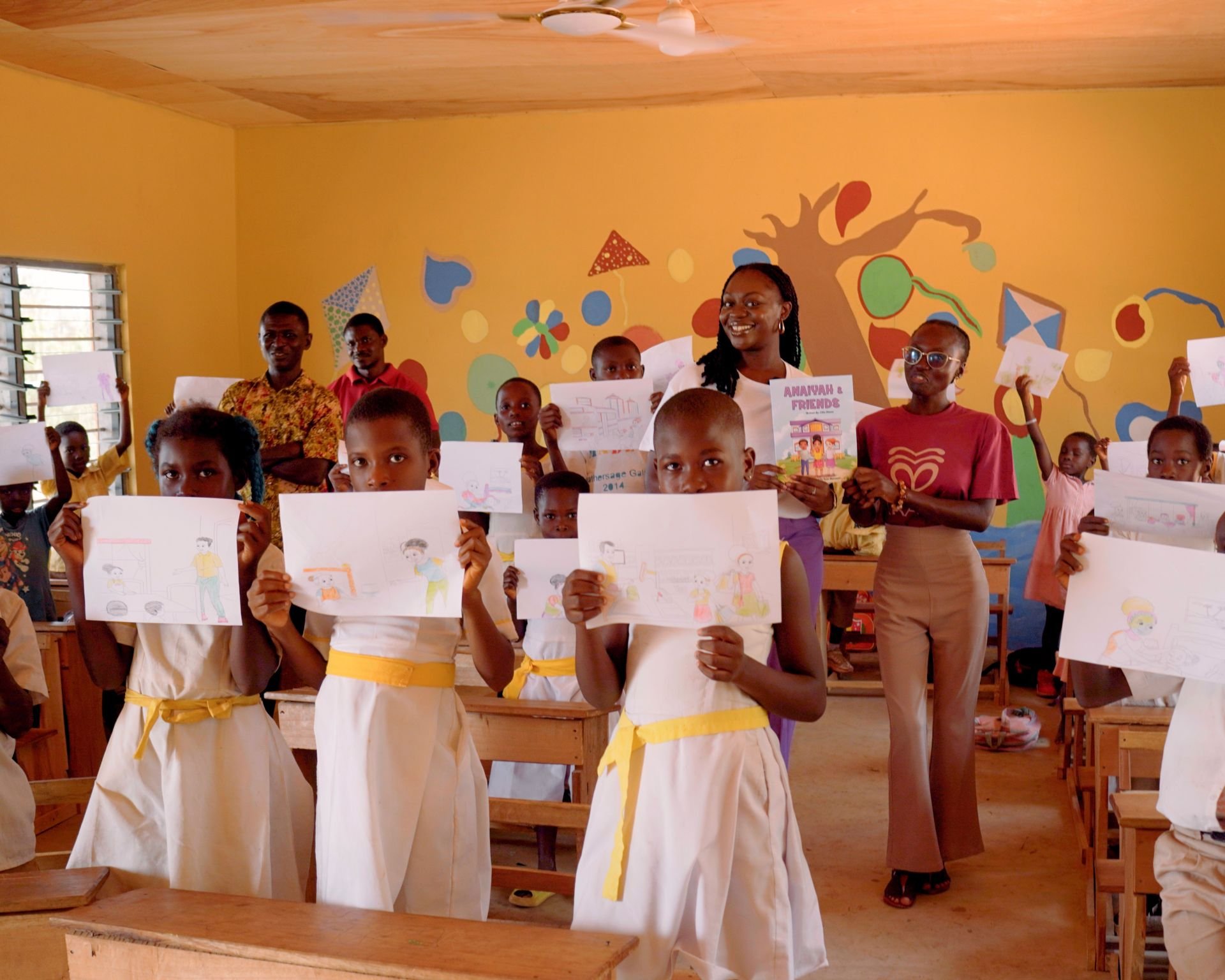 Children and teachers in a classroom holding up drawings, with a colorful mural on the yellow wall behind them, and wooden desks visible.