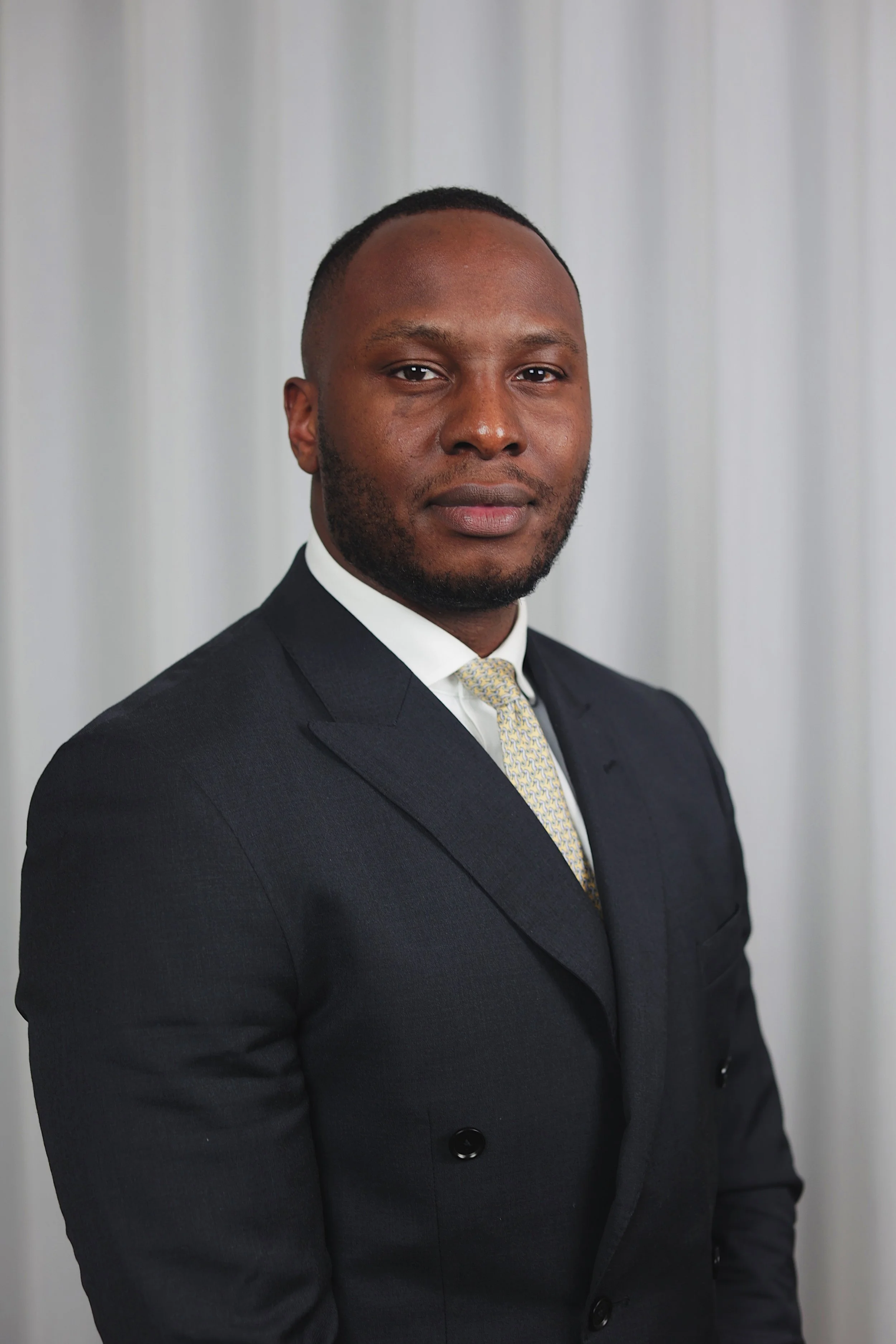 A man in a dark suit with a white shirt and patterned tie posed for a portrait against a plain, light-colored curtain background.