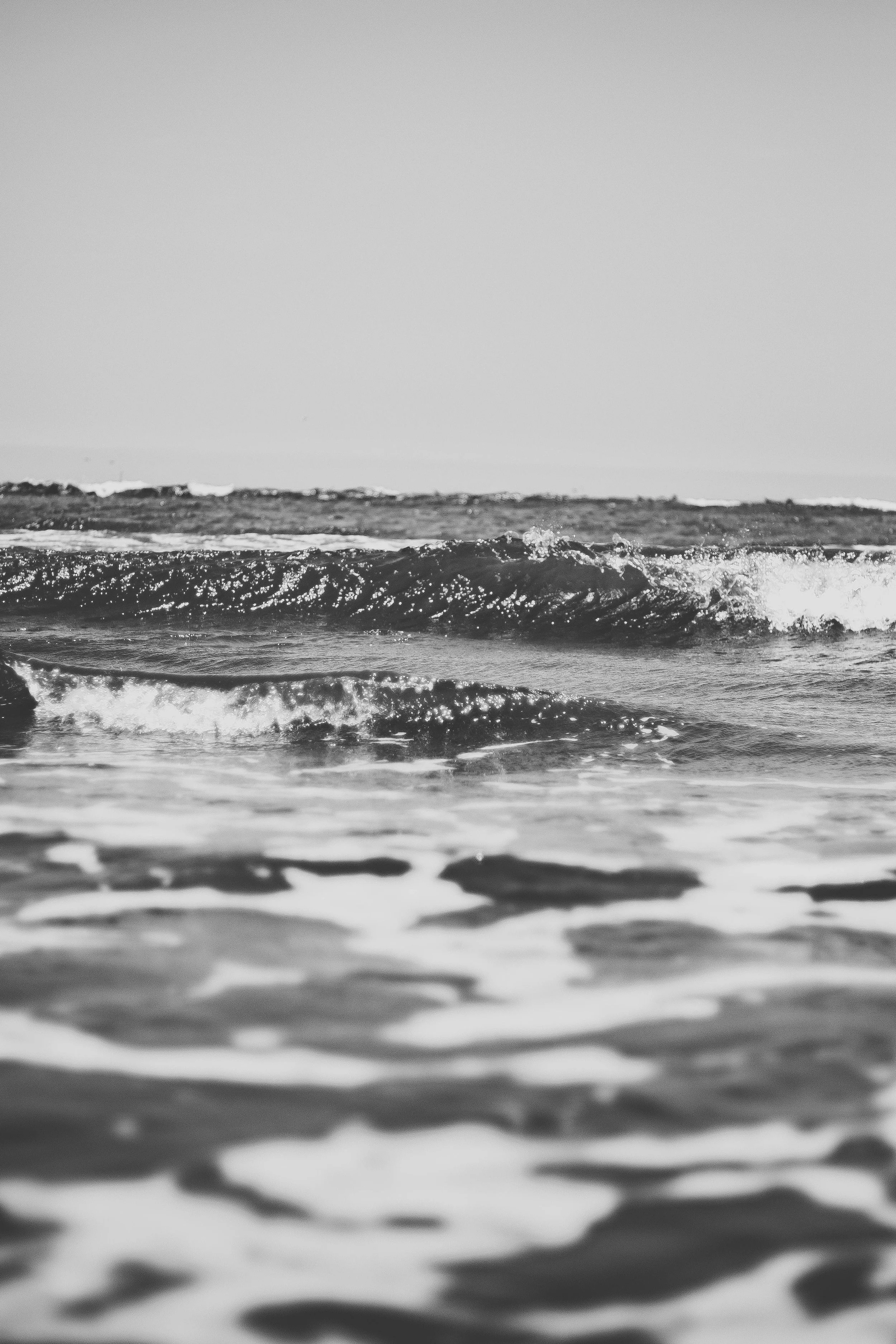 Black and white photo of ocean waves crashing near the shore with a clear sky in the background.