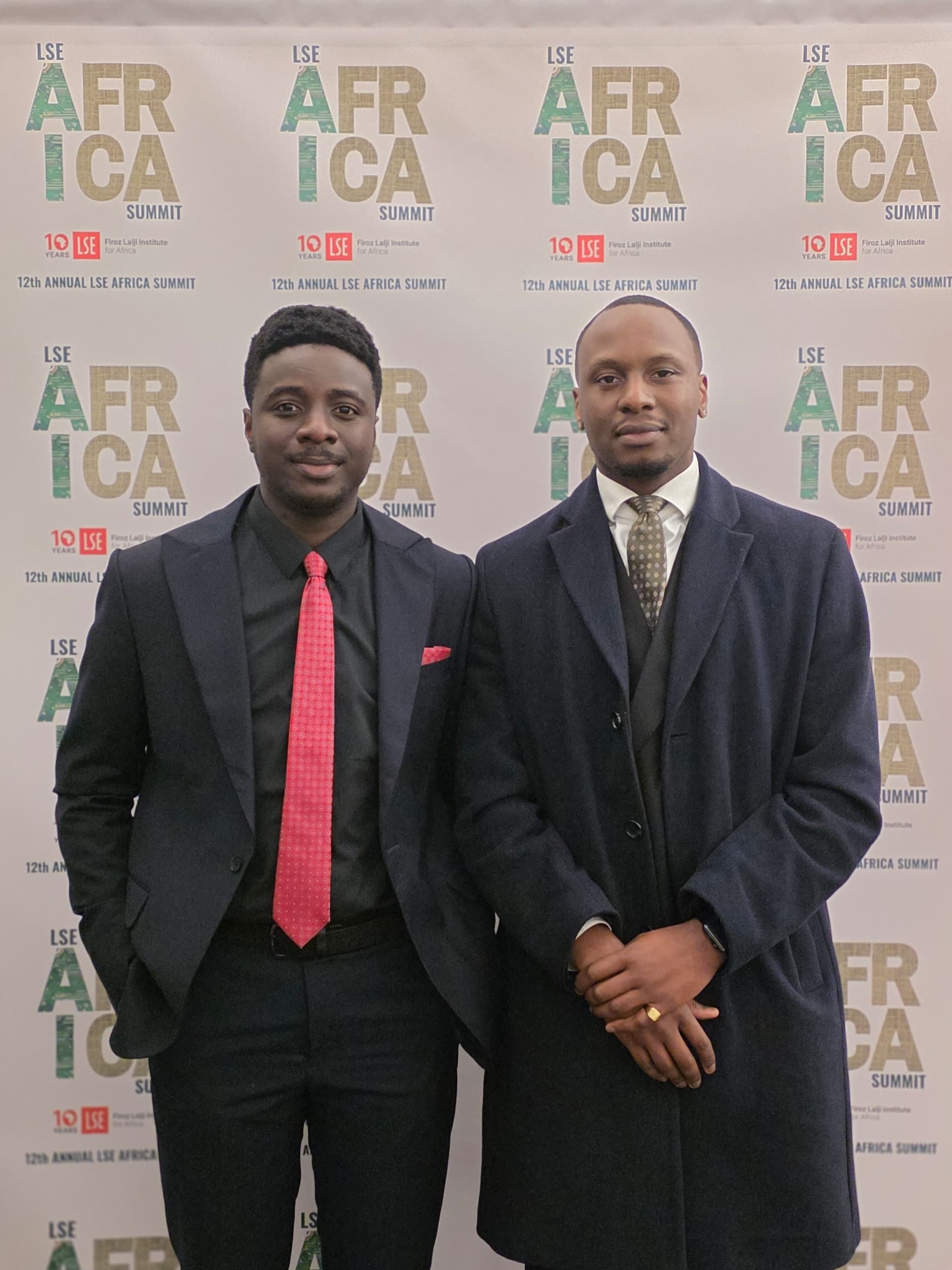 Two men in formal attire standing in front of a backdrop at the LSE Africa Summit. The man on the left wears a dark suit with a red tie and a black shirt. The man on the right wears a dark overcoat with a patterned tie and white shirt. Both are posing for the photo.