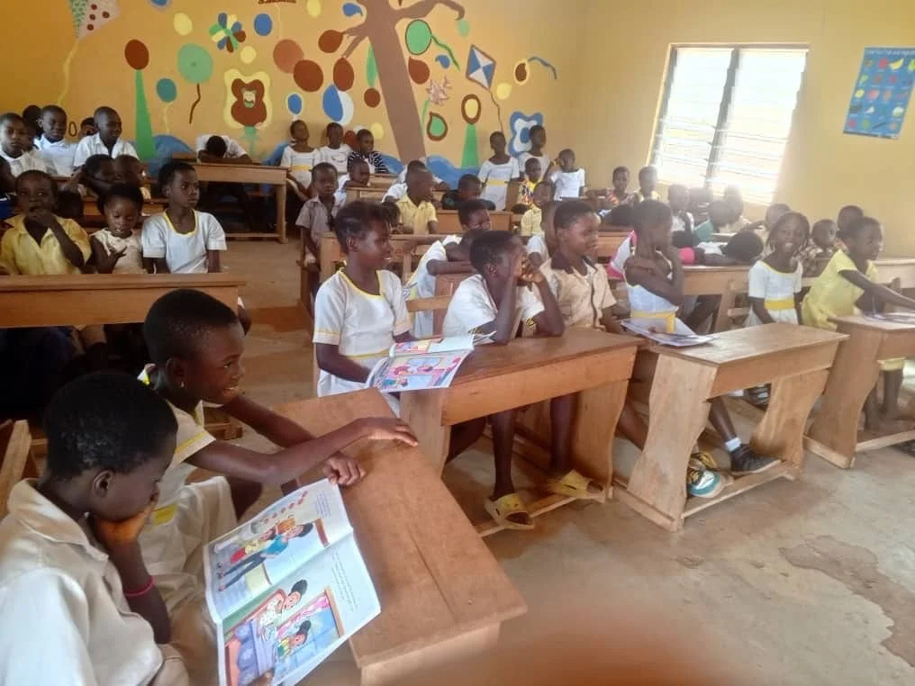 A classroom filled with young students sitting at wooden desks, reading books, with colorful drawings of animals and a tree on the yellow-painted walls, and sunlight coming through the open window.