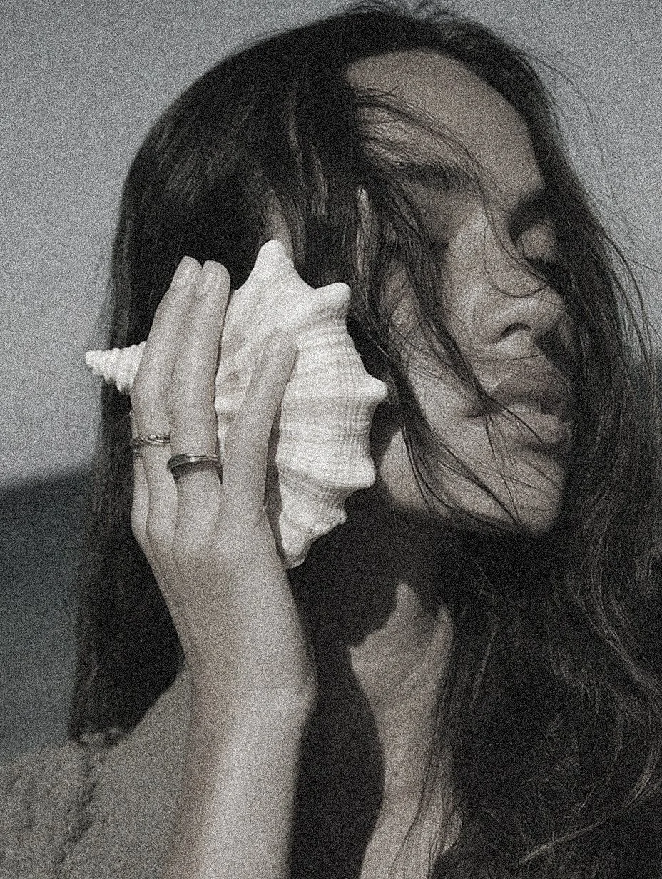 A black and white photo of a woman with long hair holding a seashell to her ear, with her eyes closed and a peaceful expression.