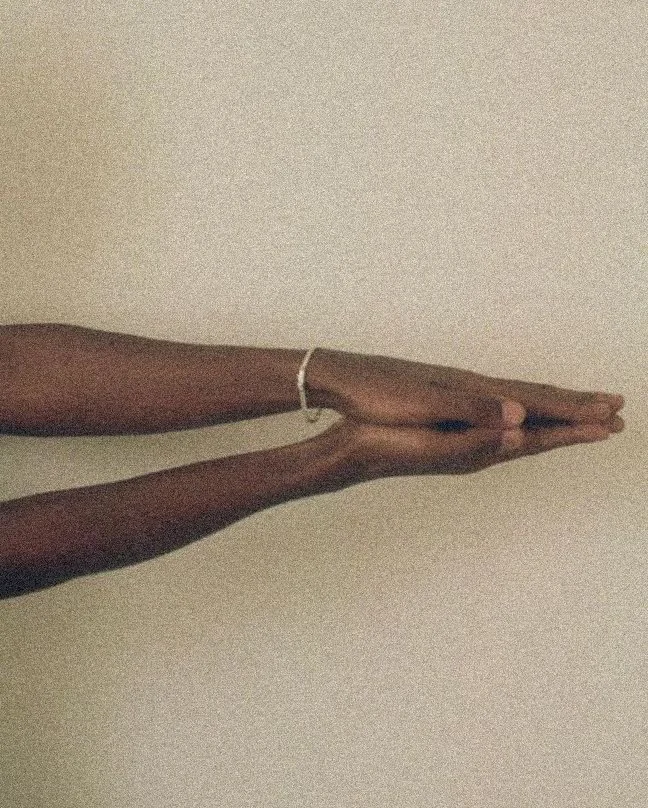 Person with dark skin extending their arms forward, palms together, against a plain light-colored wall.