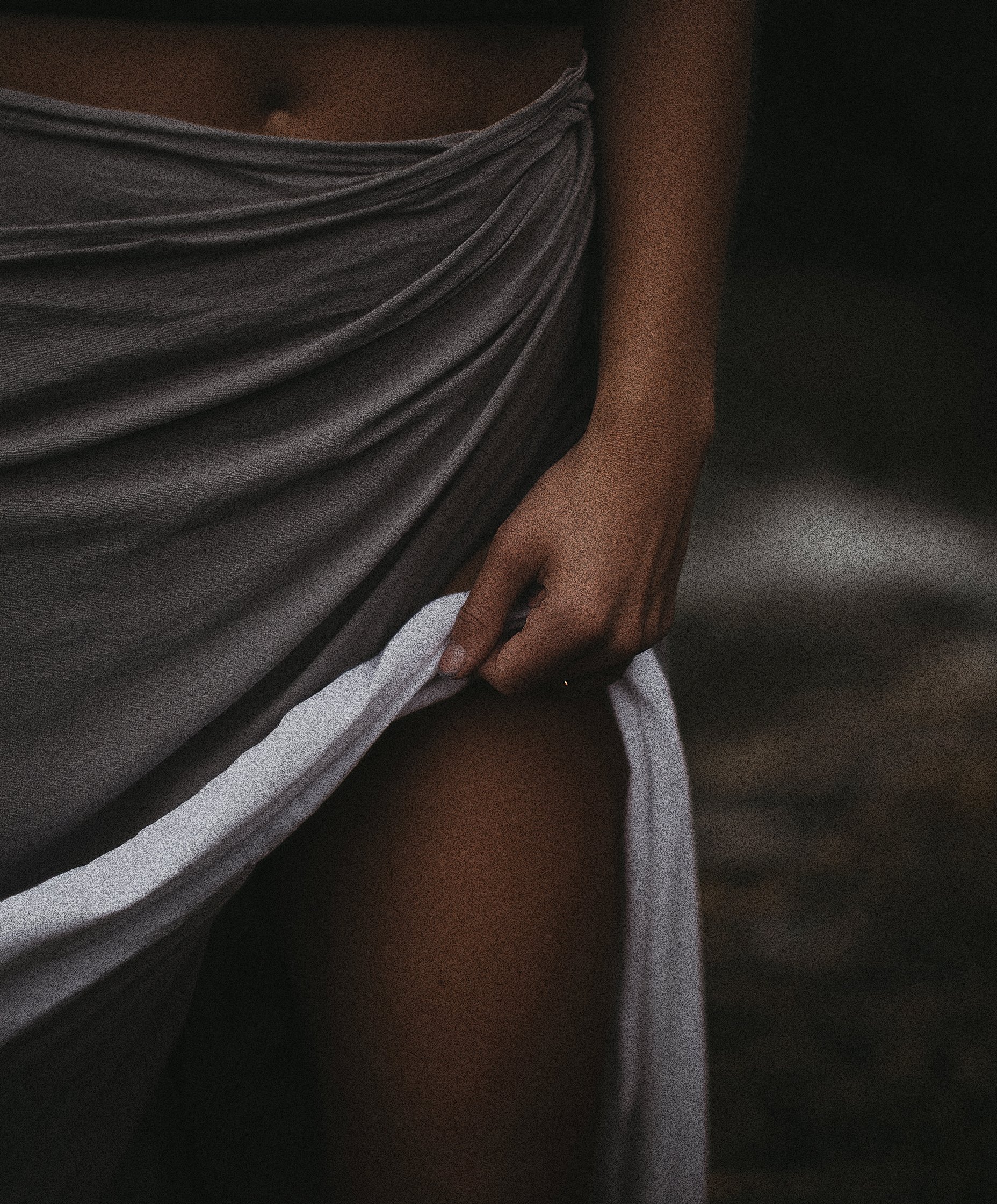A close-up photo of a person's midsection, showing a gray gathered fabric top and a brown arm resting by their side, with a dark background.