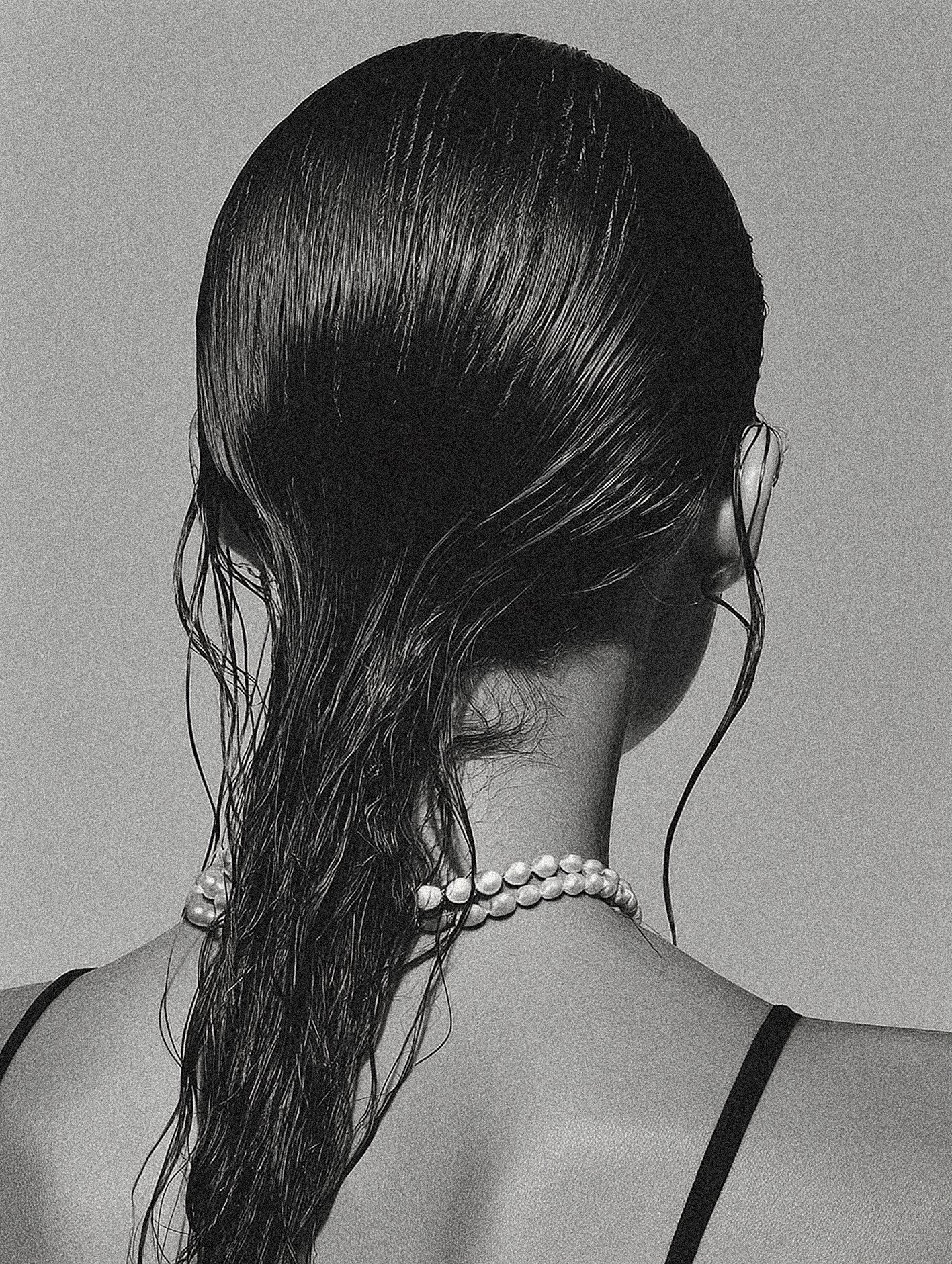 Black and white photograph of a woman's back, showing wet hair draped over her shoulder, wearing a pearl necklace and a black arm strap.