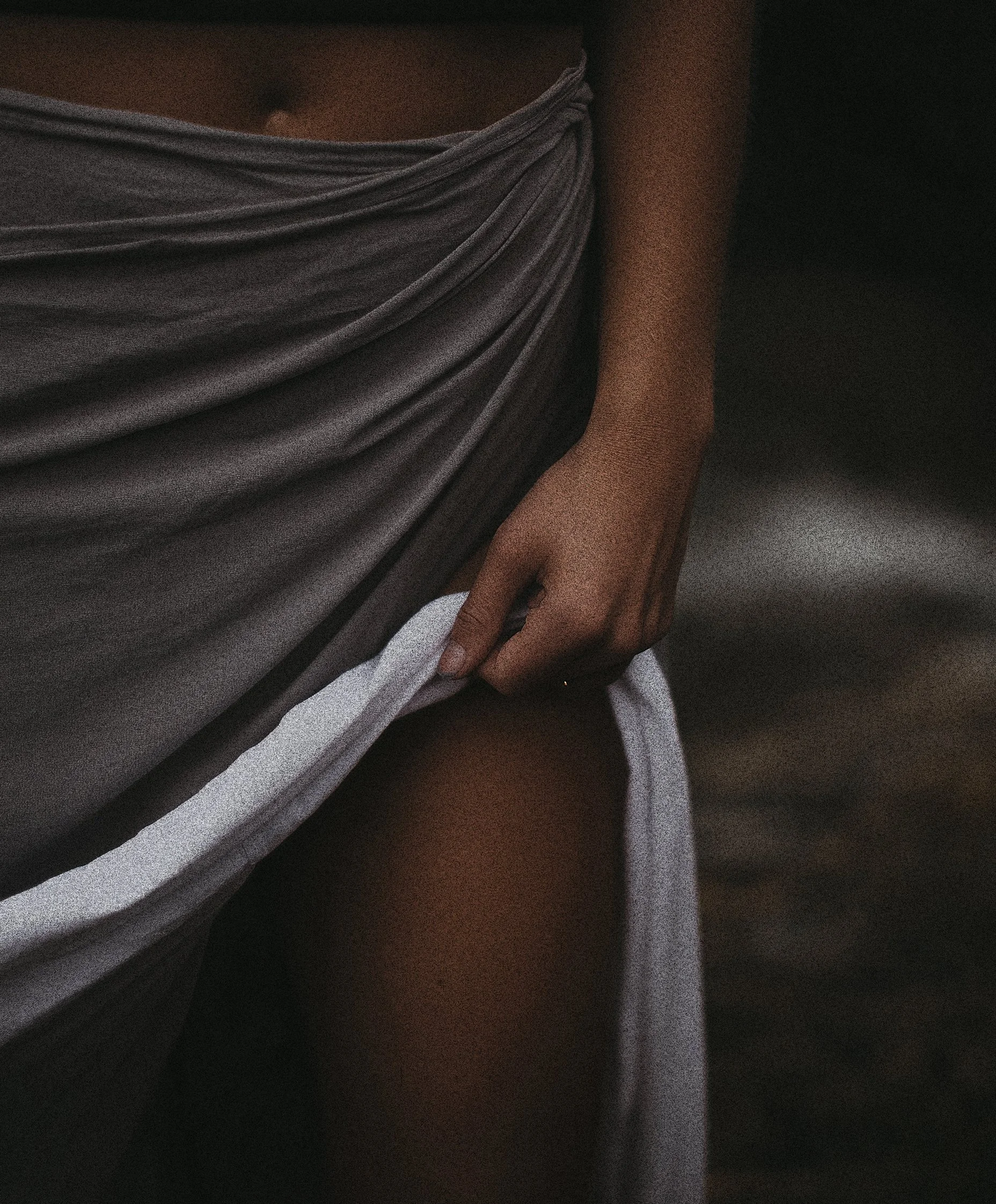 Close-up of a person's hand resting on their lap, with bare skin visible on the arm and a draped gray fabric covering part of the body. Dark background.