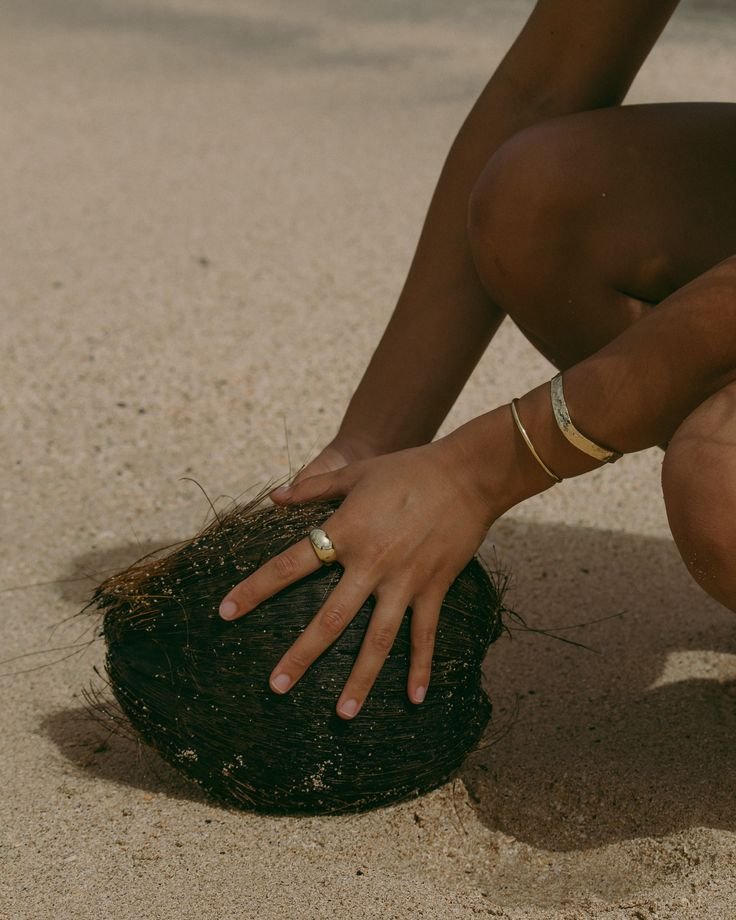 Person crouching on sandy beach with one hand on a round piece of seaweed or ocean plant and displaying rings on fingers.