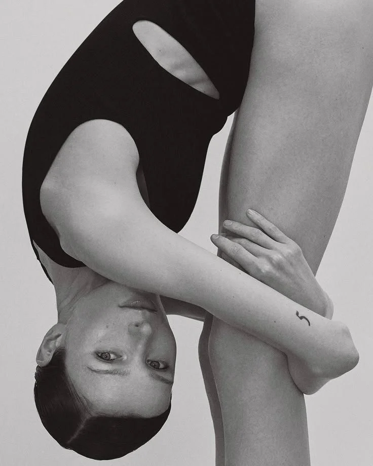 A black and white photo of a woman with short hair and a piercing on her eyebrow, lying upside down with her head near the bottom of the image. She is wearing a black top with cutouts and has minimal makeup. Her left arm is wrapped around her leg, and she has a small tattoo on her wrist.