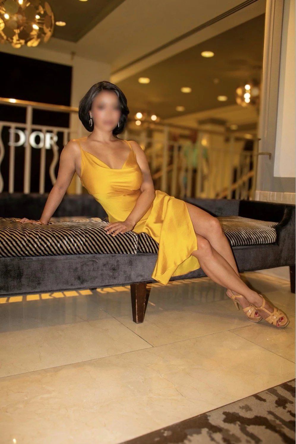 A woman in a yellow satin dress sitting on a lounge chair in an indoor setting, with a blurred background of a hotel or lounge area.