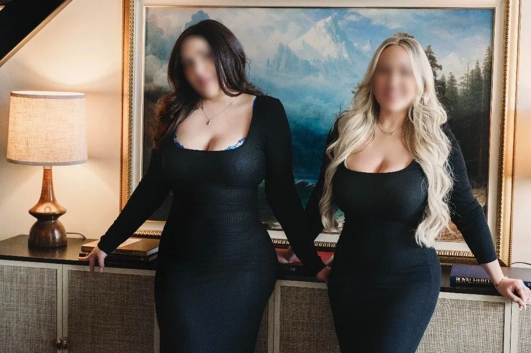 Two women in black dresses standing side by side in front of a landscape painting, with a lamp on a side table to the left.