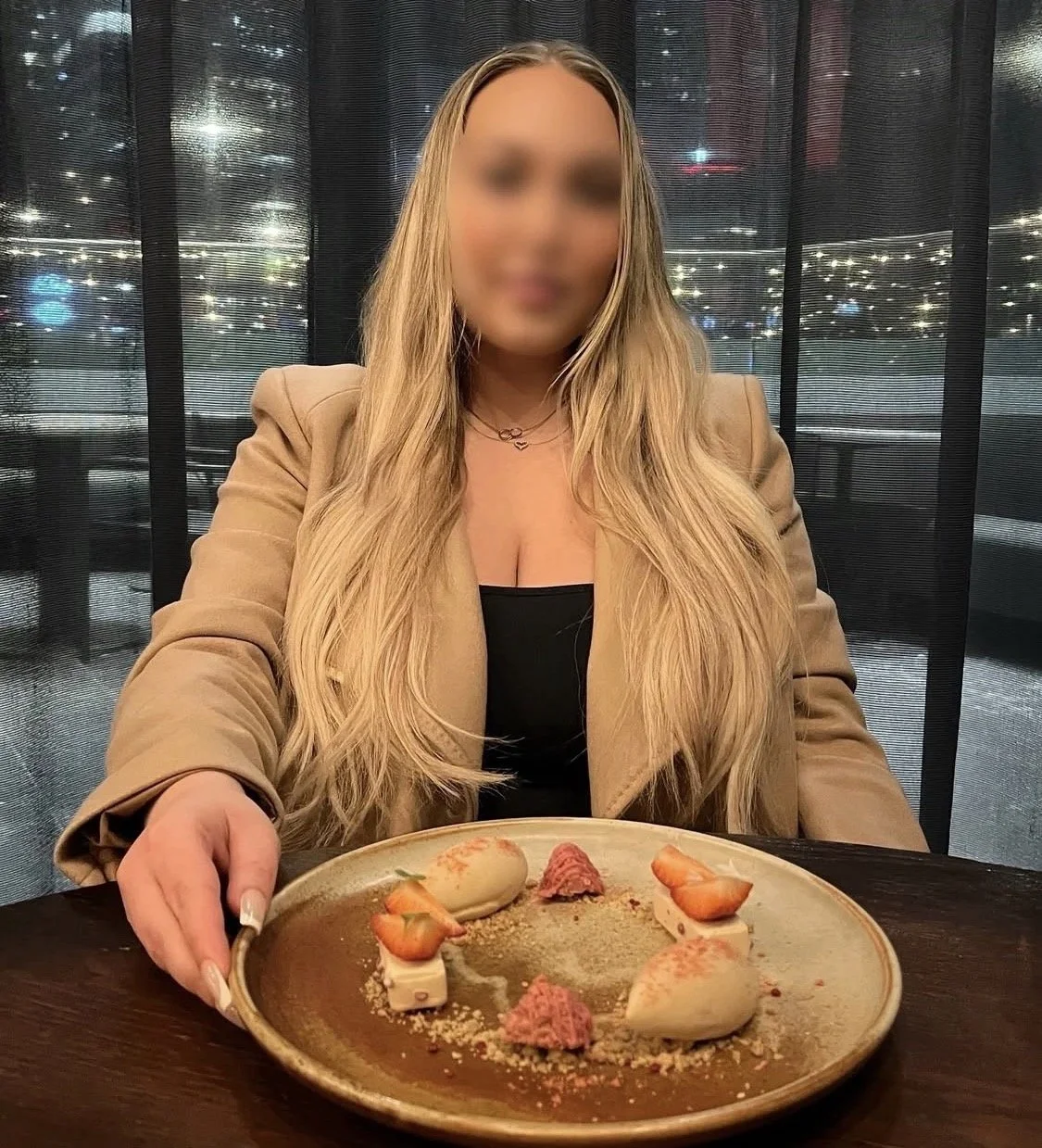 A woman with long blonde hair sitting at a table in a restaurant, holding a plate with dessert. The background shows a dark window with city lights outside.