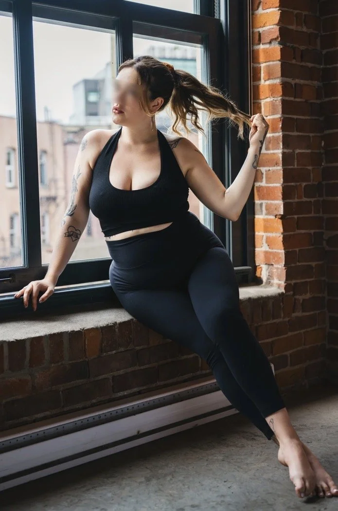 A woman in black athletic wear sitting on a window sill in a studio with brick walls. She is holding her ponytail and looking out the window.