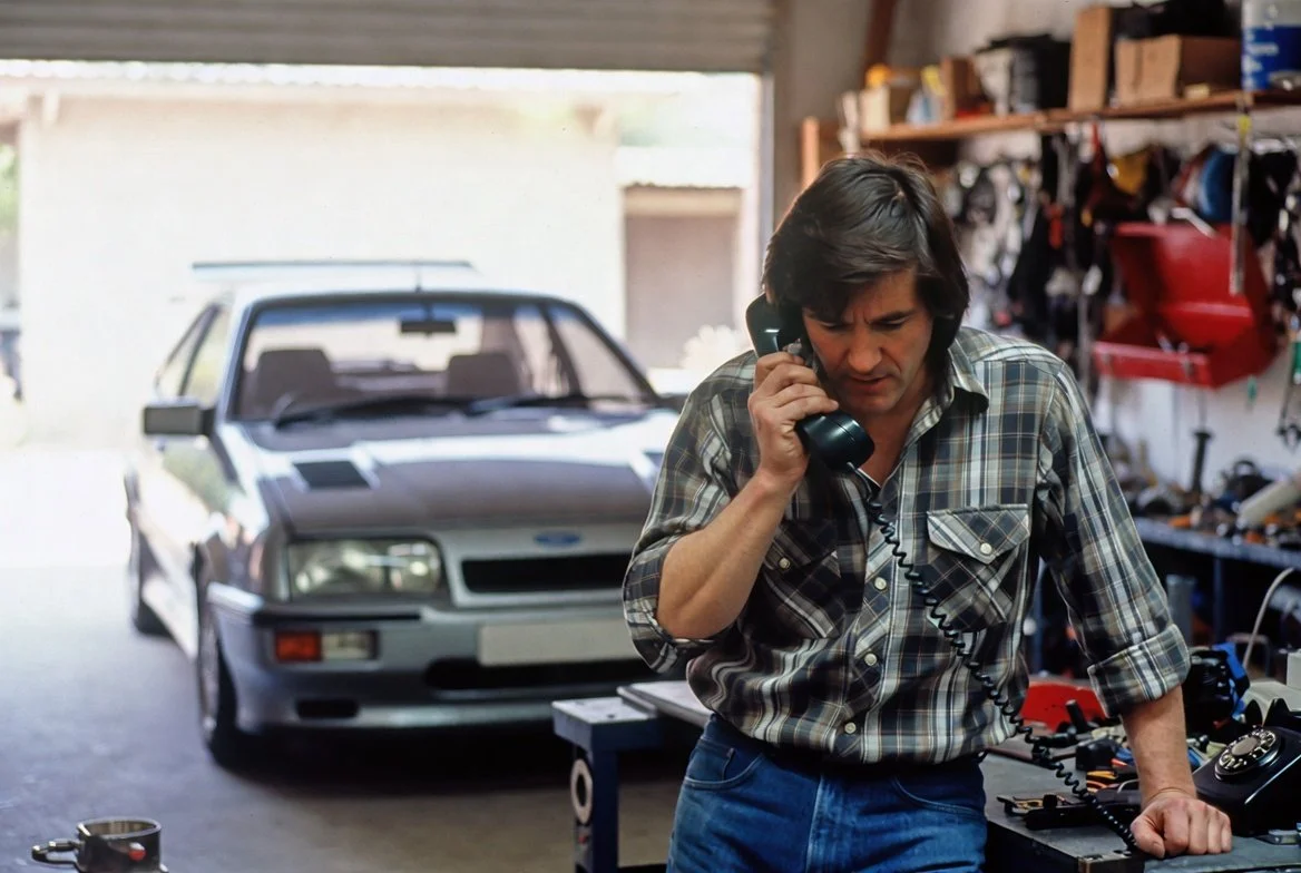 Man with dark hair wearing a plaid shirt, talking on a landline phone inside a garage with a silver car in the background and shelves with tools and supplies.