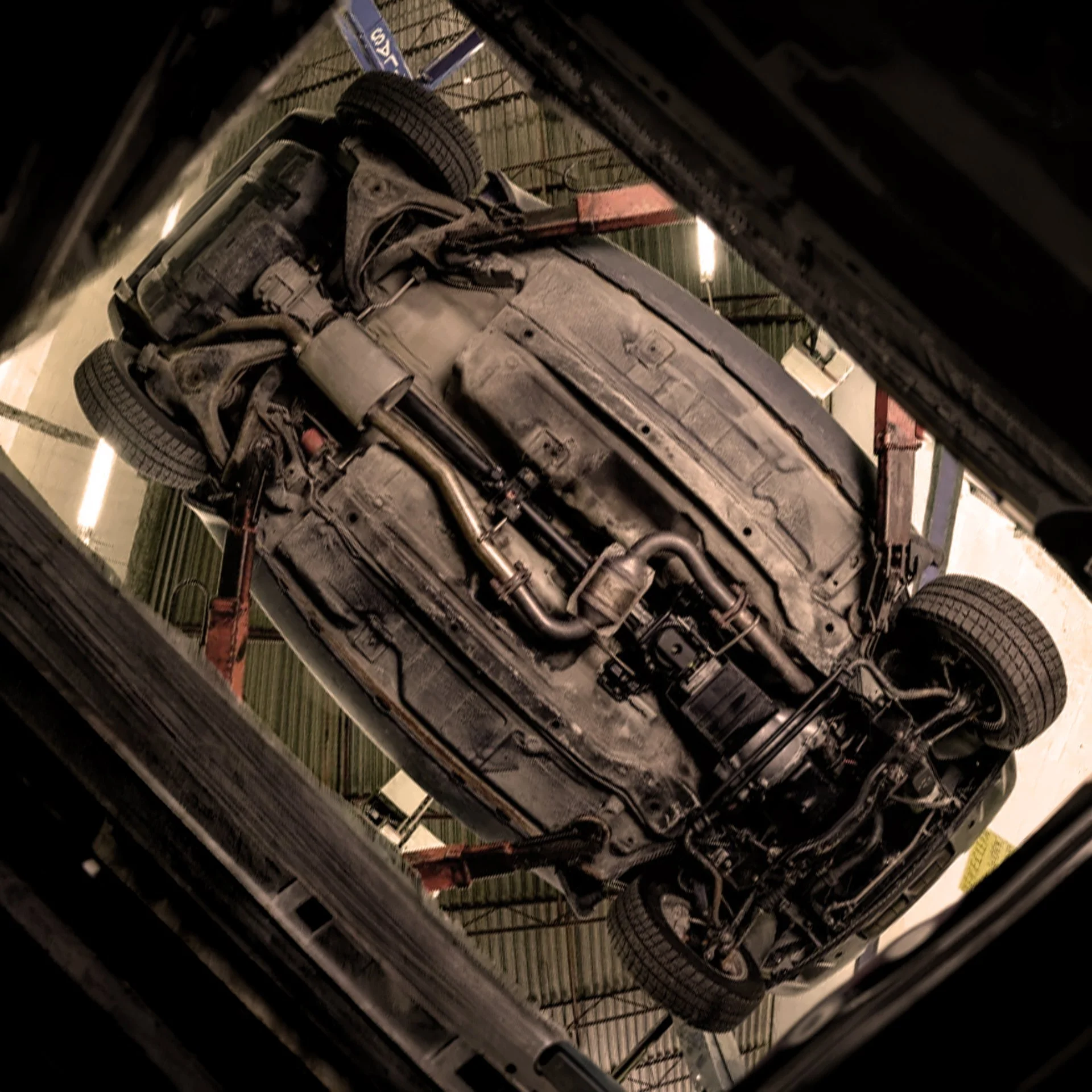 Underneath view of a dusty black car flipped upside down on a hydraulic lift in a garage.
